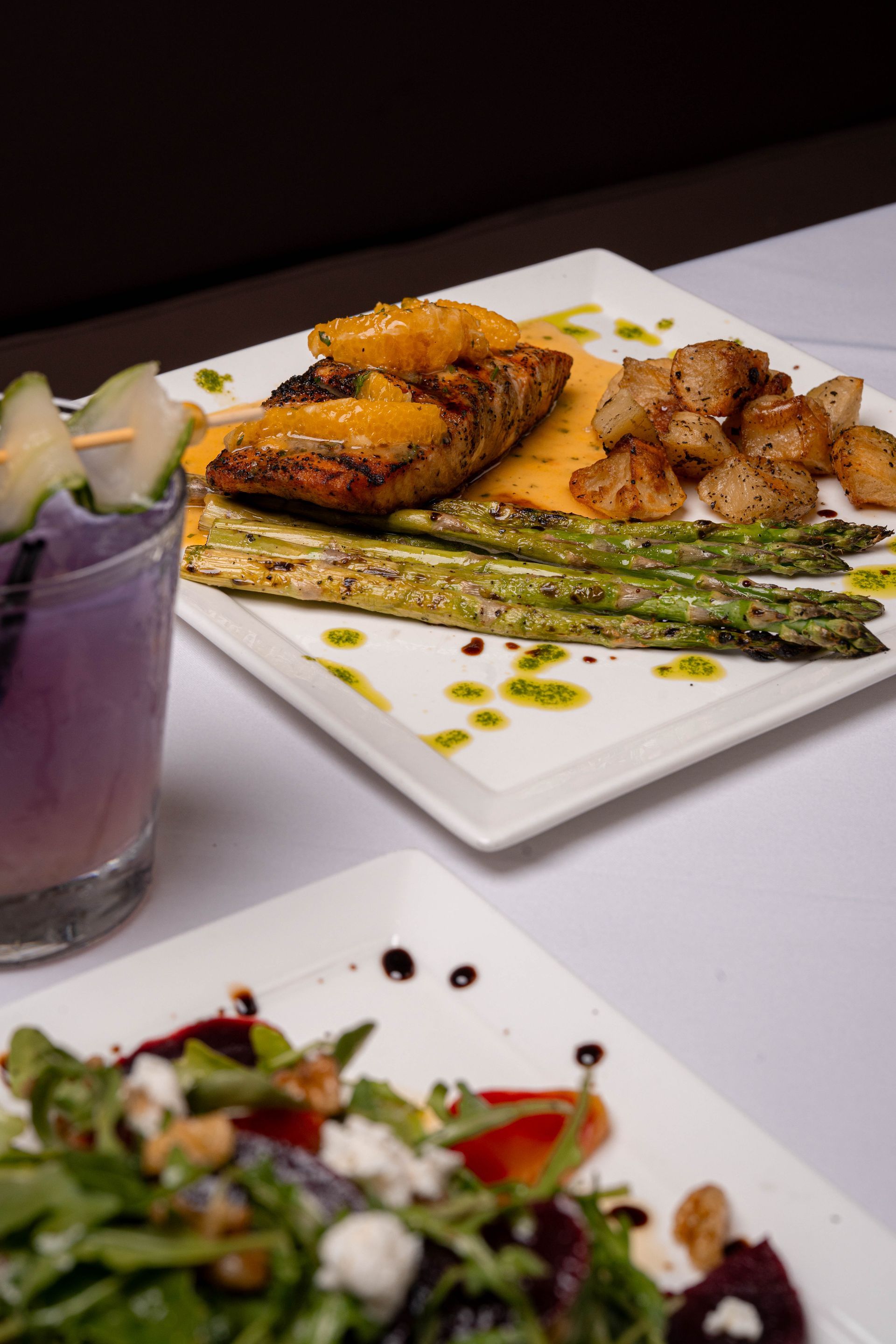 Grilled salmon with asparagus and potatoes, a purple cocktail, and a salad on a table with a white tablecloth.