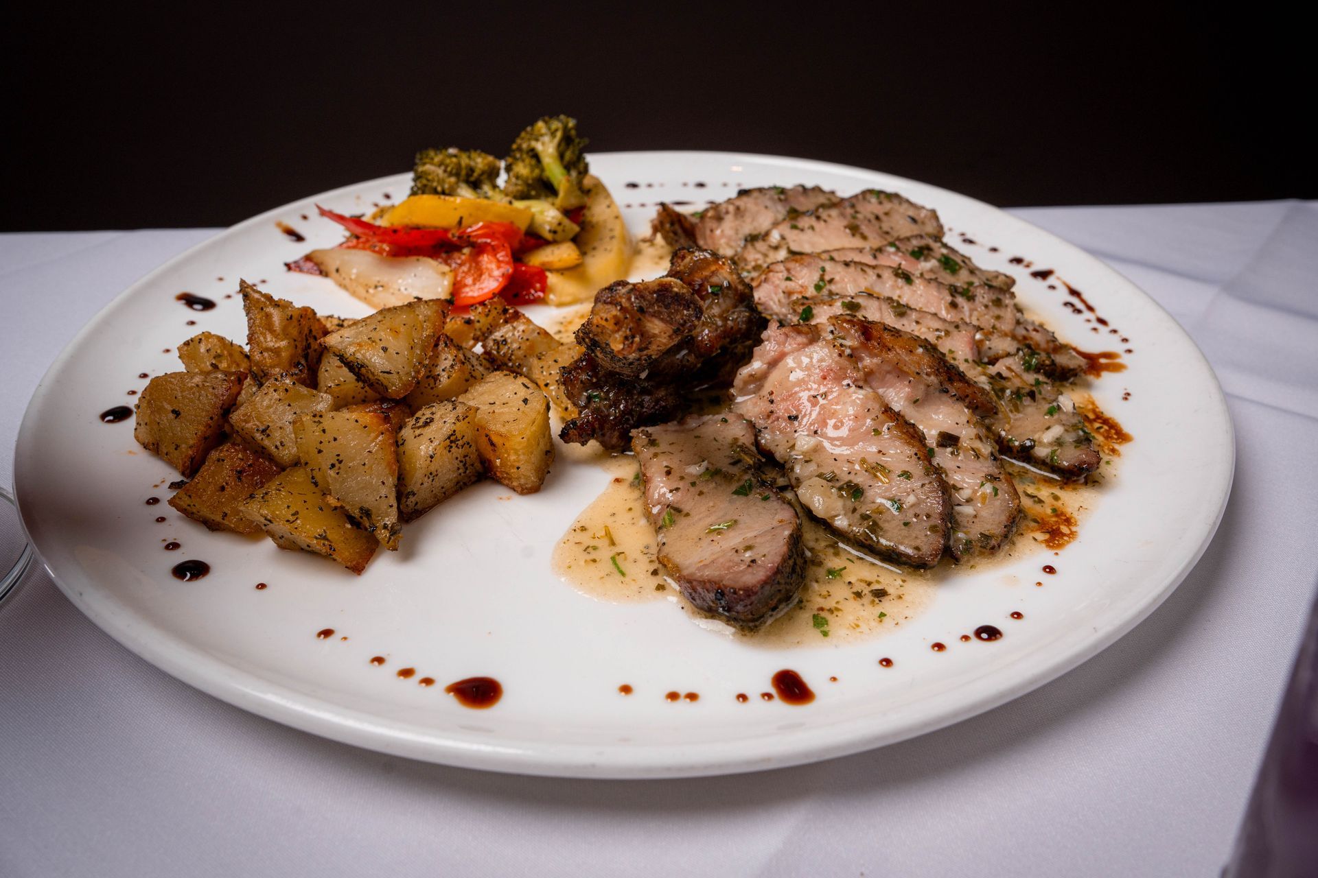 White plate with roasted potatoes, mixed vegetables, meat, and sauce. On a white tablecloth with a dark background.