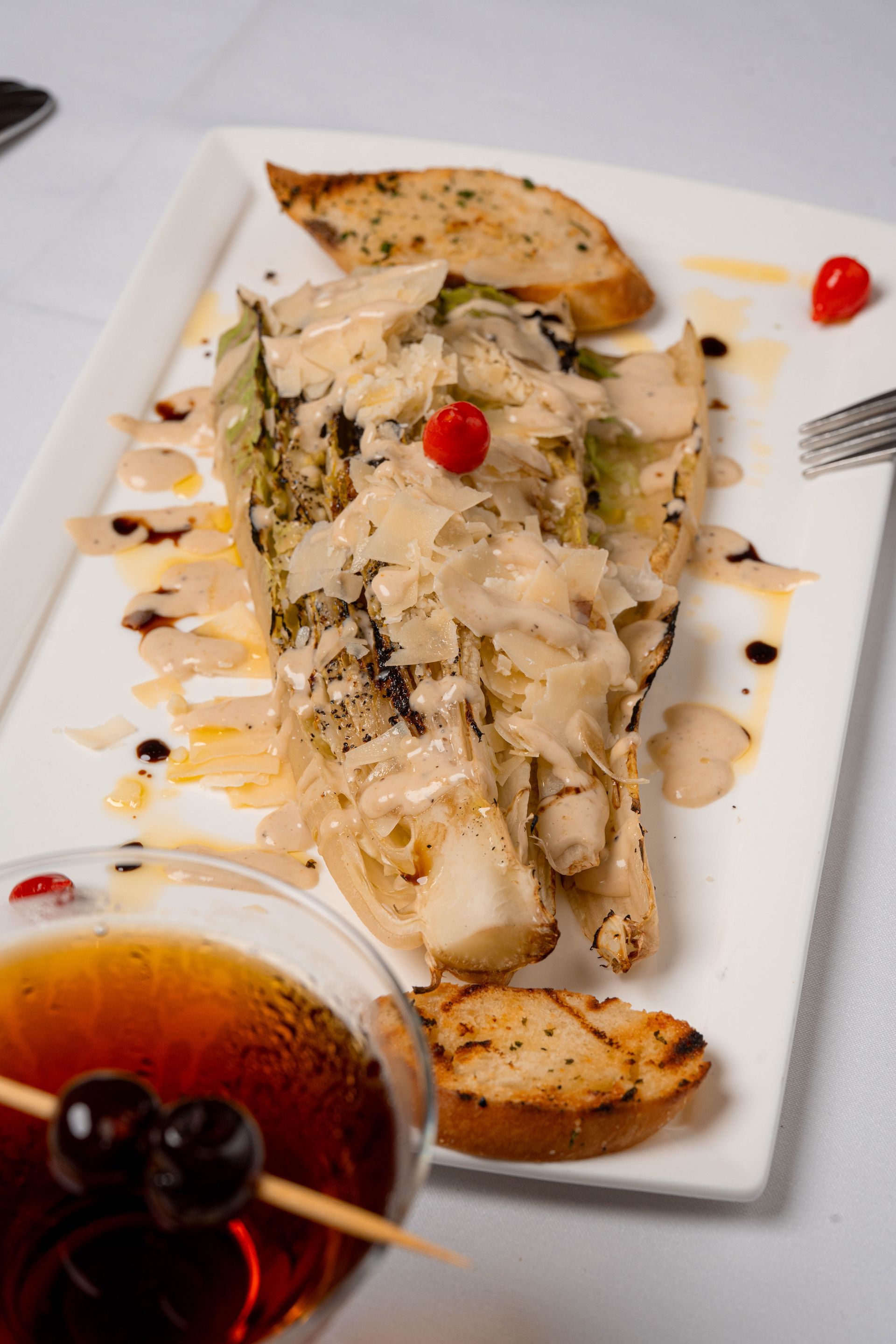 Grilled romaine salad with shaved cheese, dressing, and toast points, on a white plate, next to a cocktail with cherries.