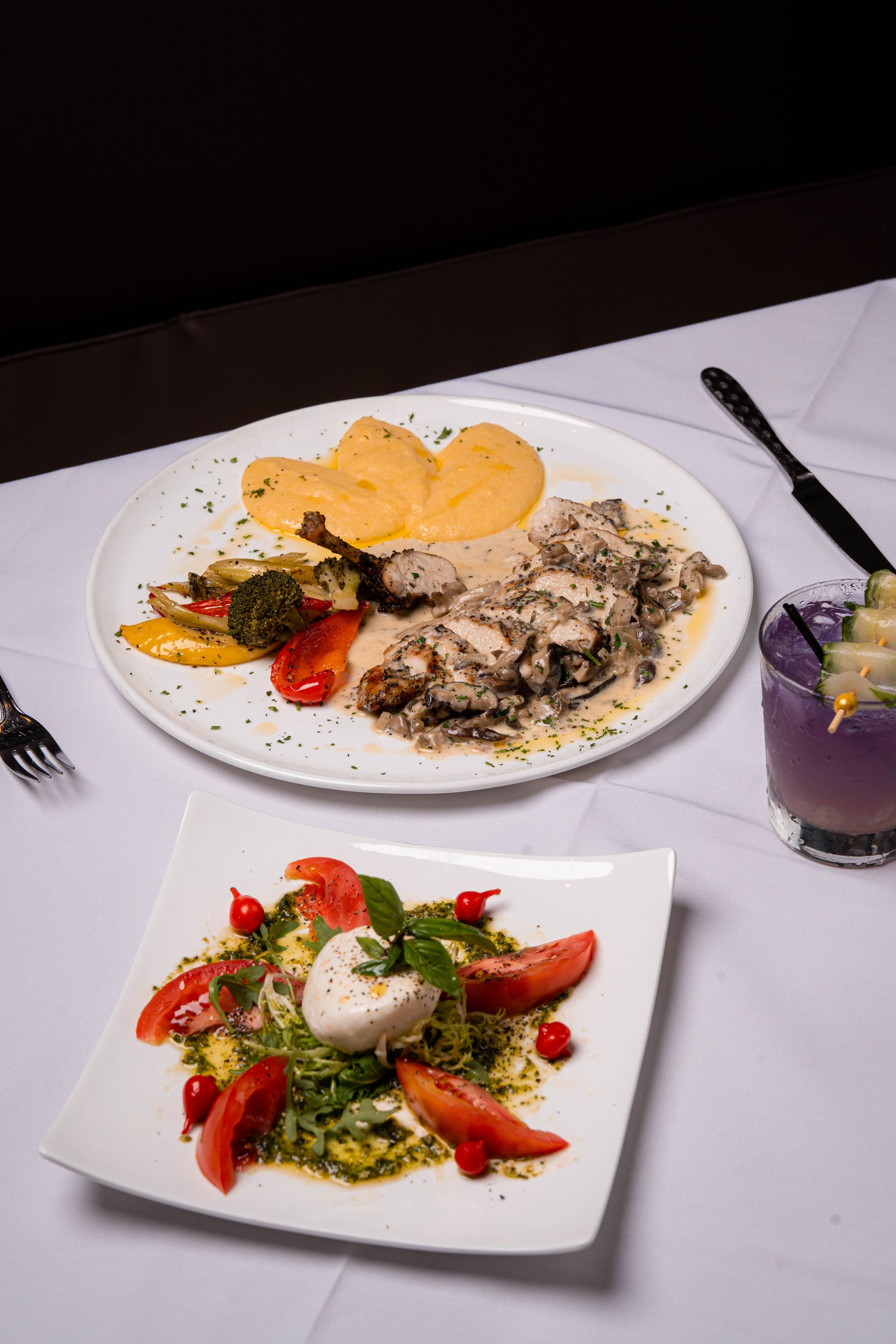 Two plates of food on a white tablecloth. Top plate: meat with sauce, potato and vegetables. Bottom plate: salad with mozzarella and tomatoes. A cocktail is in the background.