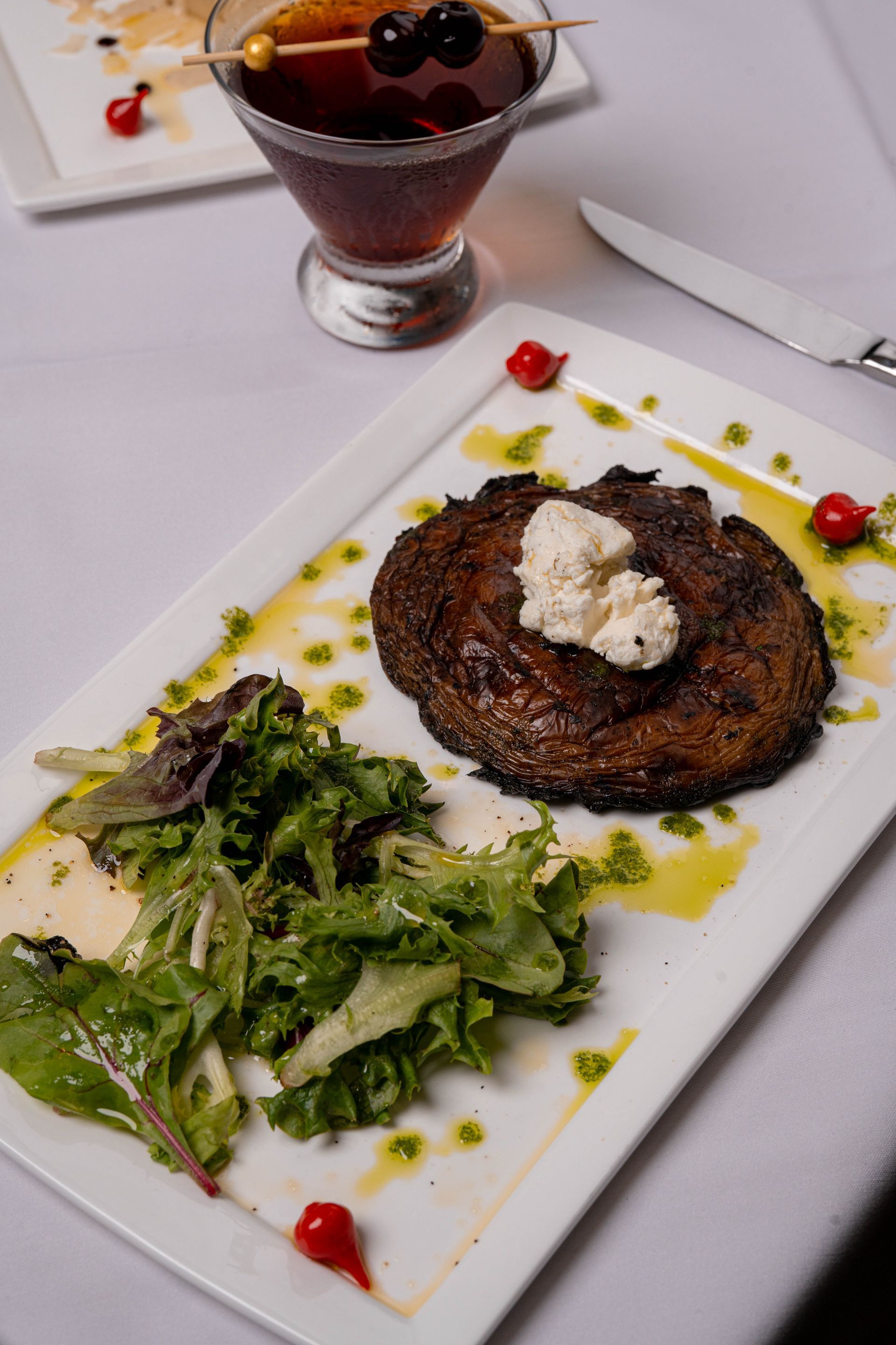 Steak with a dollop of white cream, green salad, and drink with cherries on a white plate, on a white tablecloth.