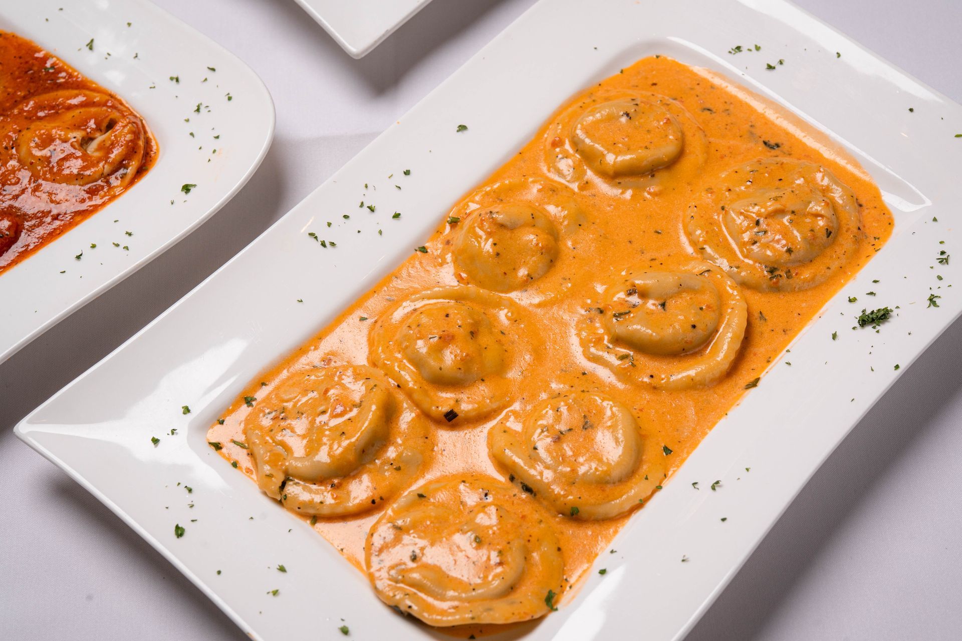 Ravioli in creamy, orange sauce on a white rectangular plate, garnished with herbs. Another plate of pasta on the left.