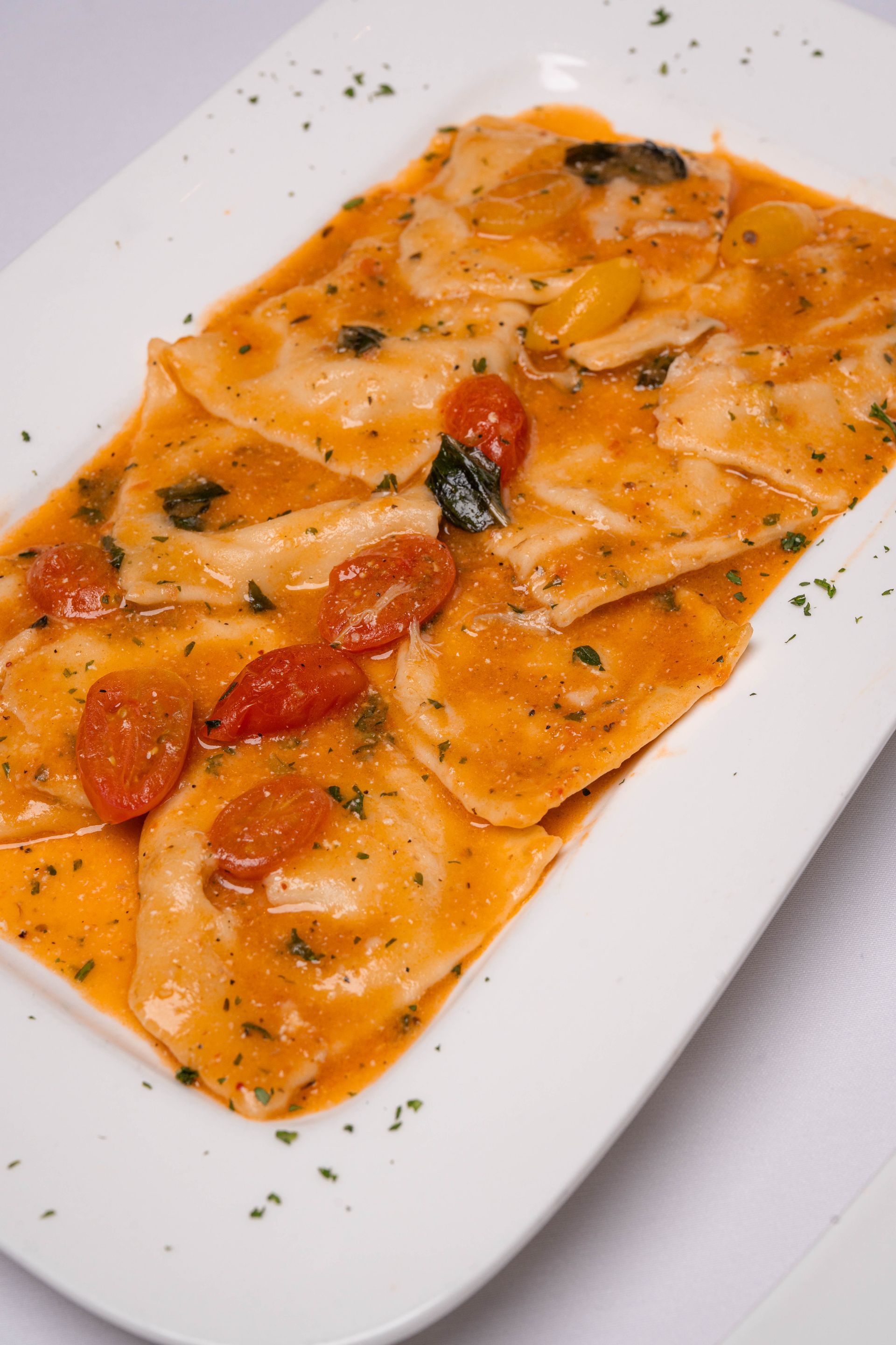 Ravioli in creamy tomato sauce, garnished with cherry tomatoes and herbs, served on a white rectangular plate.