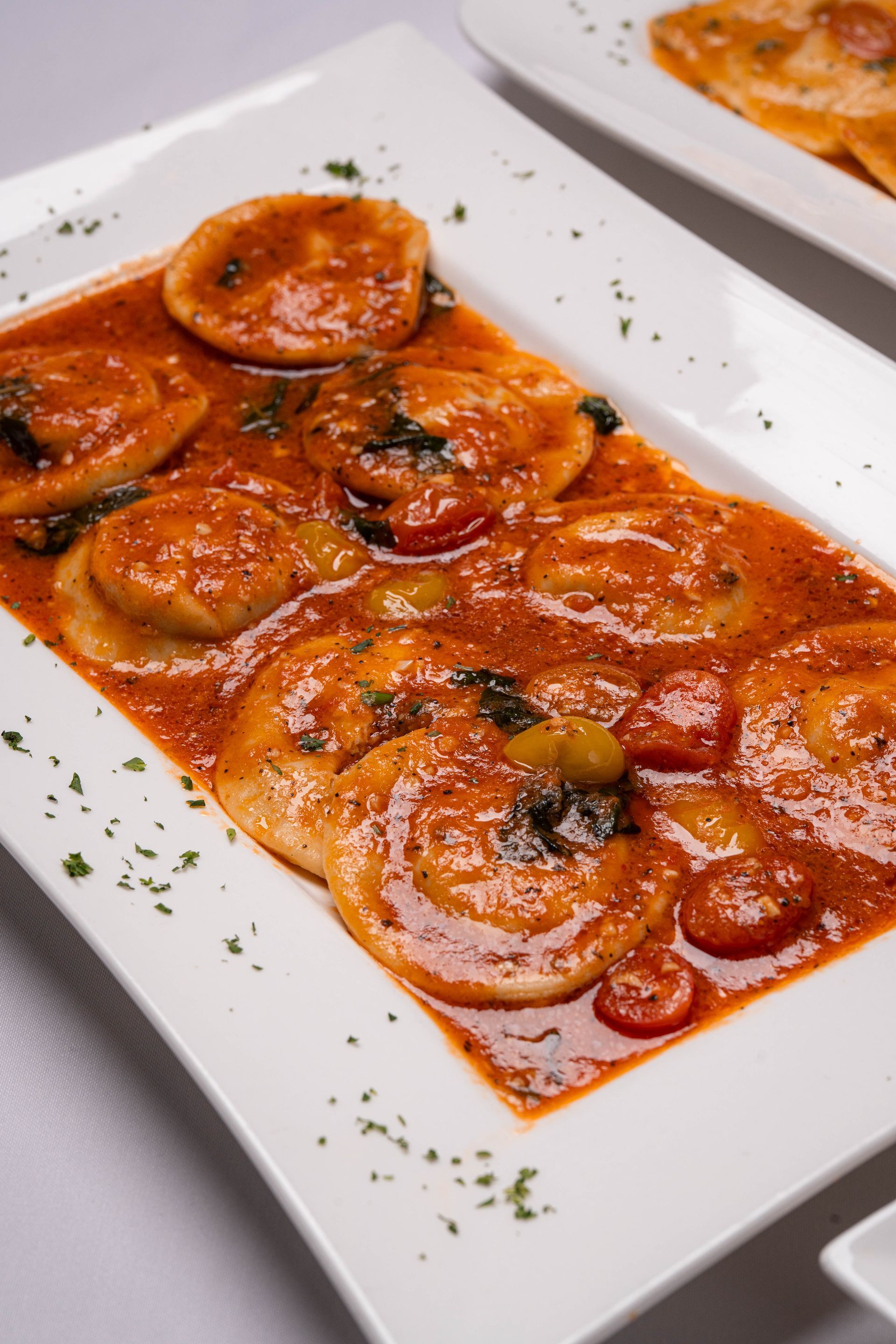 Ravioli in a red sauce, garnished with herbs, served on a white rectangular plate.