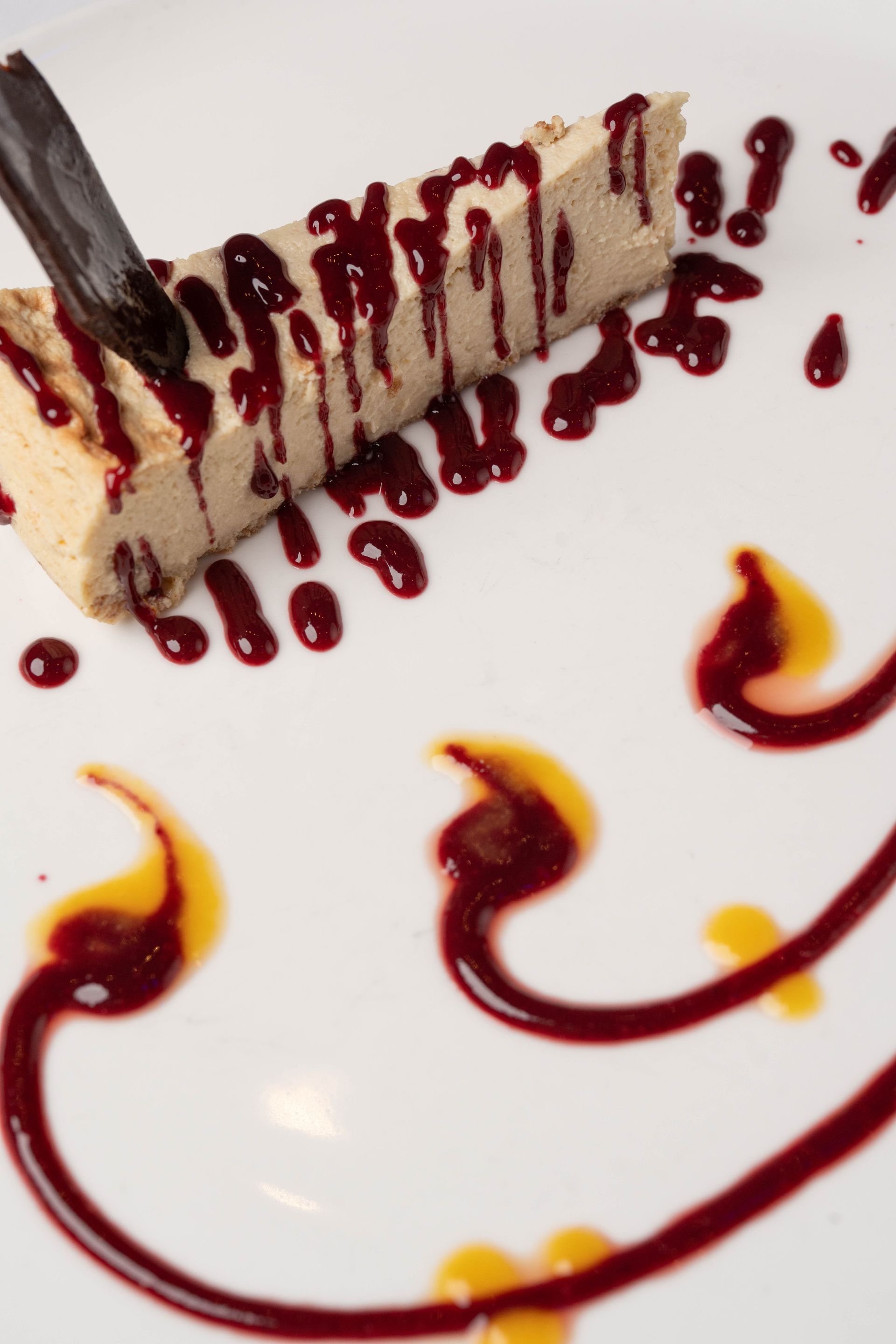 Slice of dessert with red berry sauce drizzled over, with decorative swirls of sauce on a white plate.