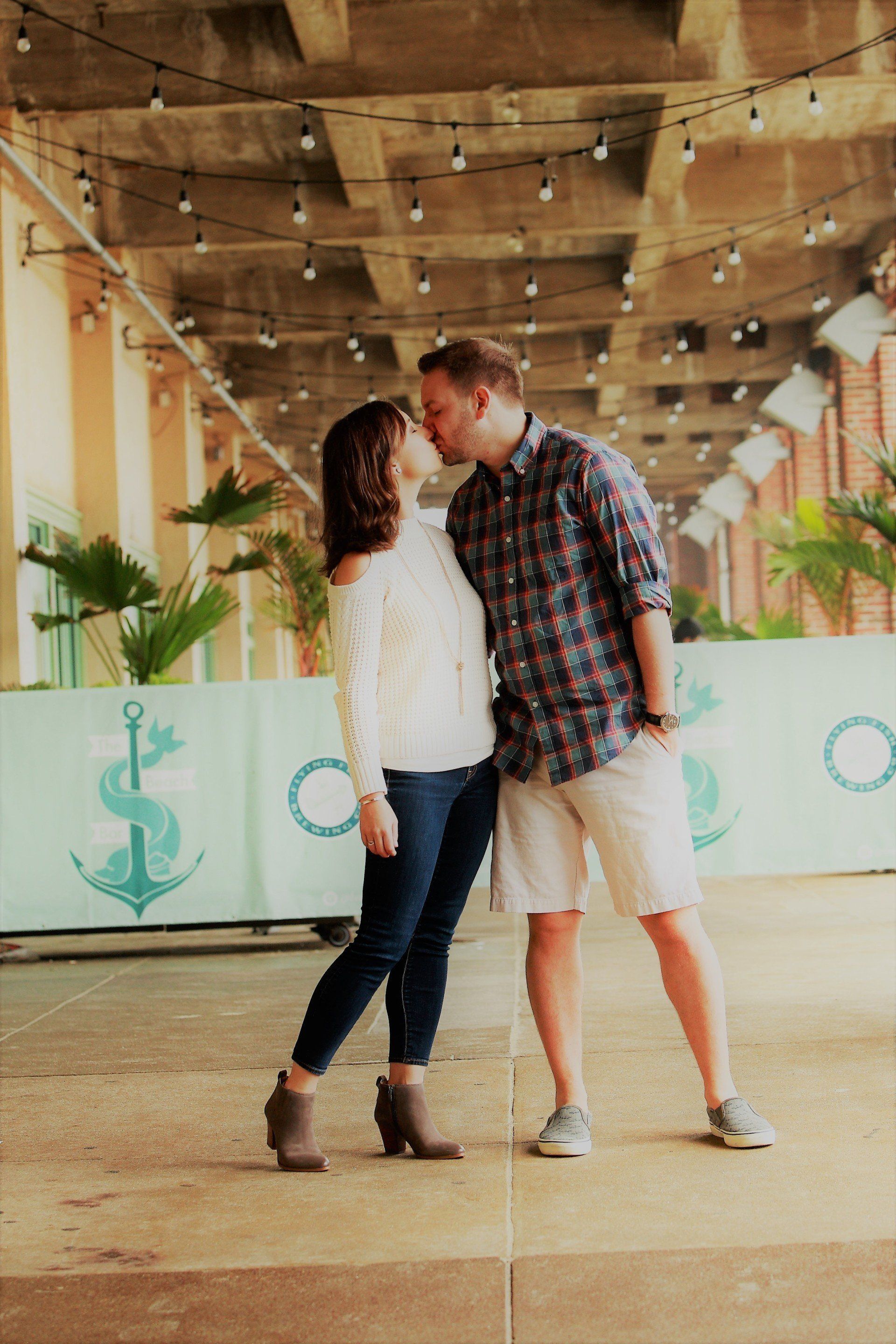 Couple kissing under a structure with string lights, nautical mural behind them.