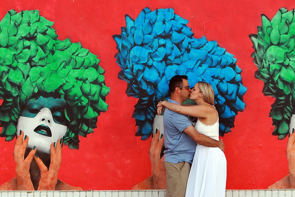 Couple kissing in front of a colorful mural of faces with blue and green hair against a red background.