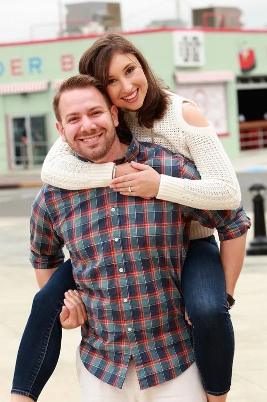 Man giving woman a piggyback ride, both smiling. Outdoors near a building with sign.