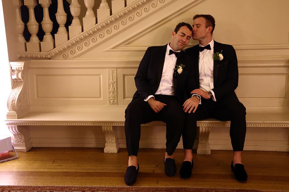 Two men in tuxedos, one kissing the other's head while seated on a bench in a building with a white staircase.