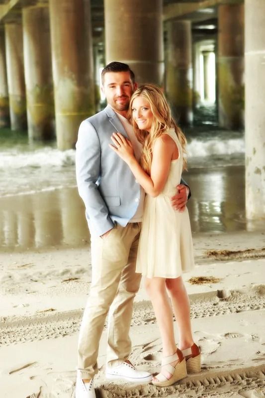 Couple embracing under a pier. Man in light blue blazer and khakis, woman in cream dress and wedges.