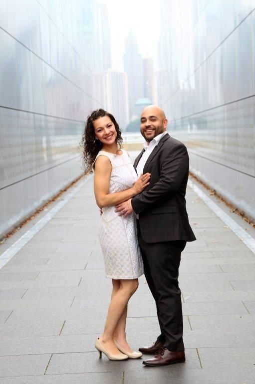 Couple smiling, embracing outdoors in front of a reflective building. Woman in white dress, man in suit.