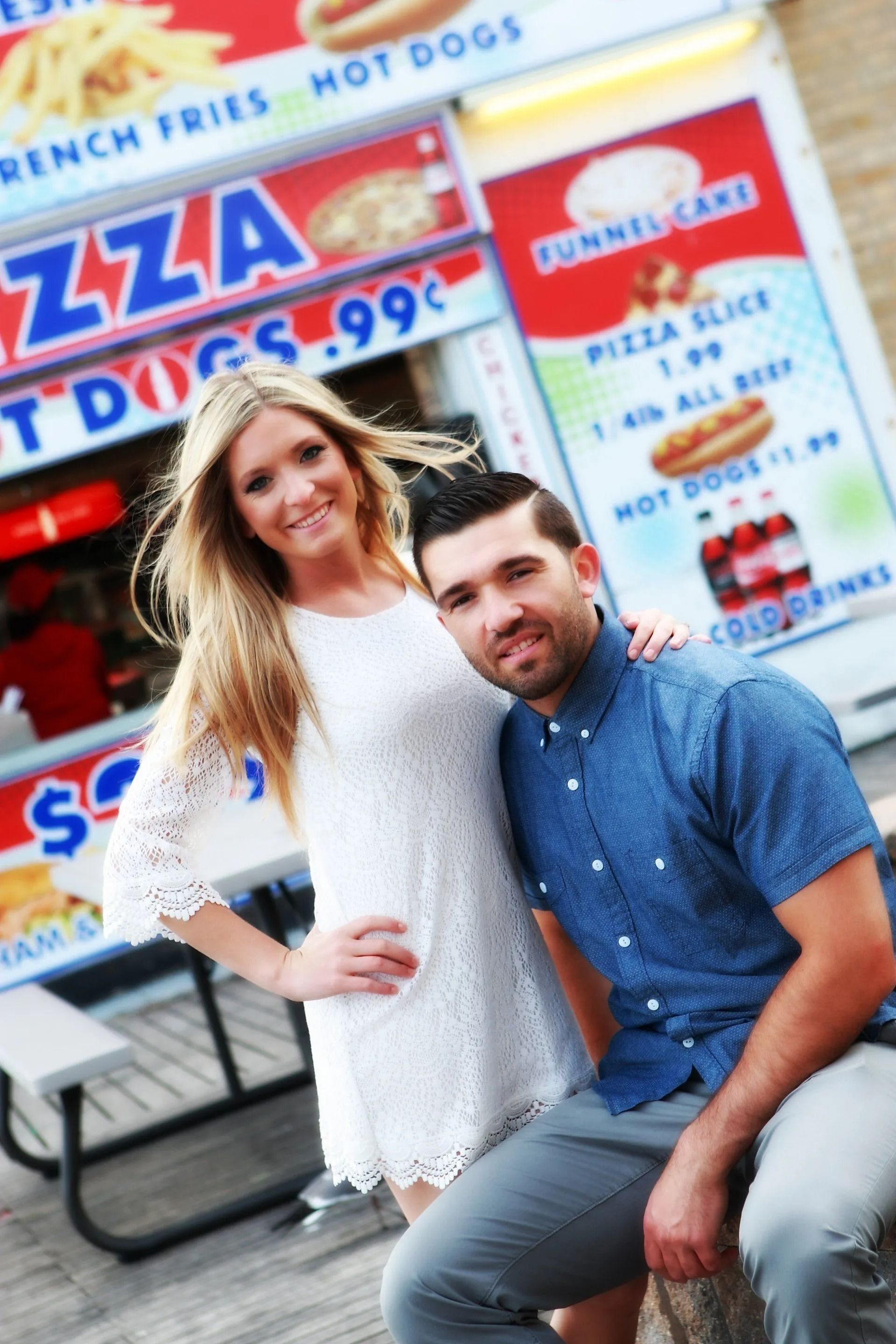 Couple posing in front of a fast food stand with menu signs. Woman in white dress, man in blue shirt.