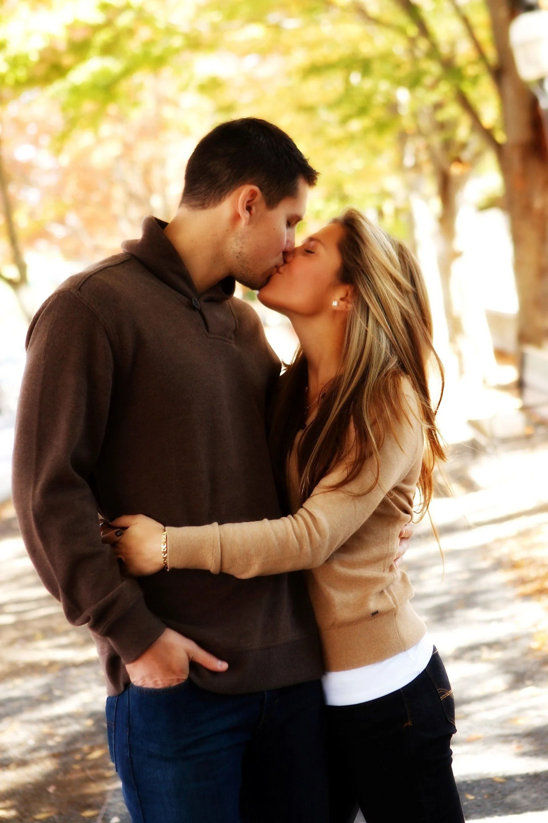 Couple kissing outdoors, man in brown sweater, woman in beige, fall foliage.