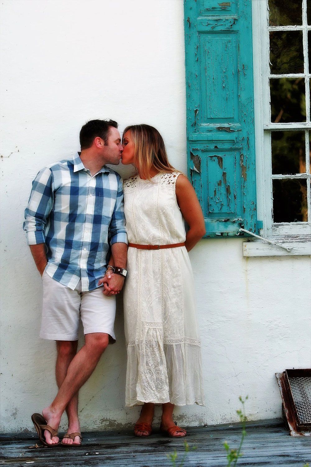 Couple kissing; standing against a white wall with a turquoise shutter; holding hands.