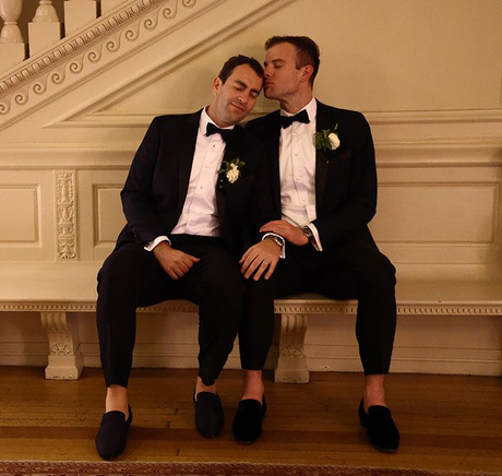 Two men in tuxedos, one kissing the other's head. They sit together on a bench indoors.