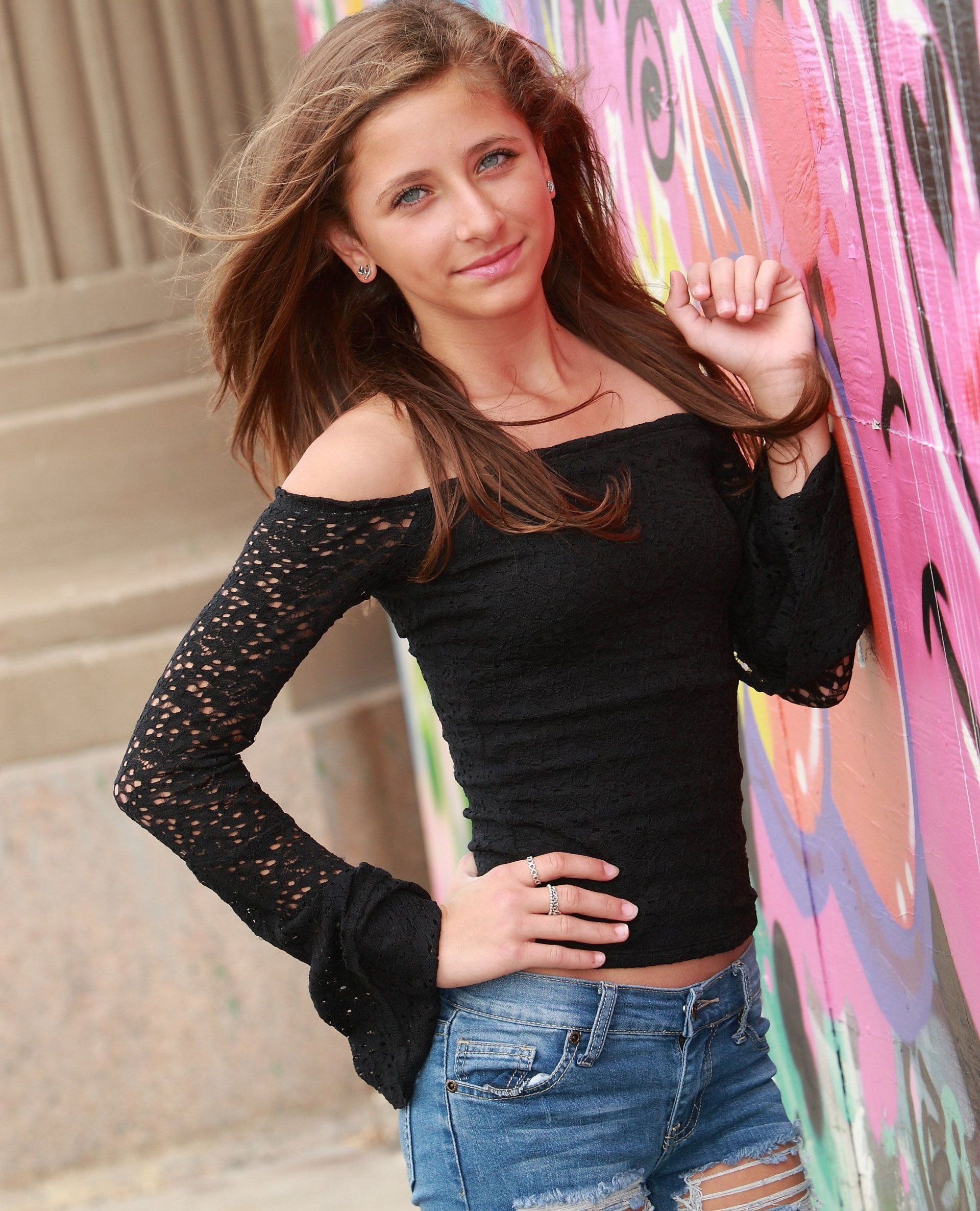 Young person in black off-the-shoulder top and ripped jean shorts leaning against a graffiti wall, smiling.