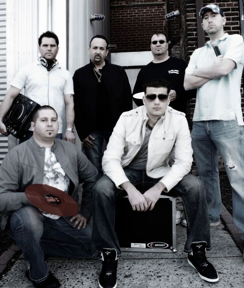 Six men pose outdoors. One holds a red record, another holds a case, and one has a device. All wear casual clothing.