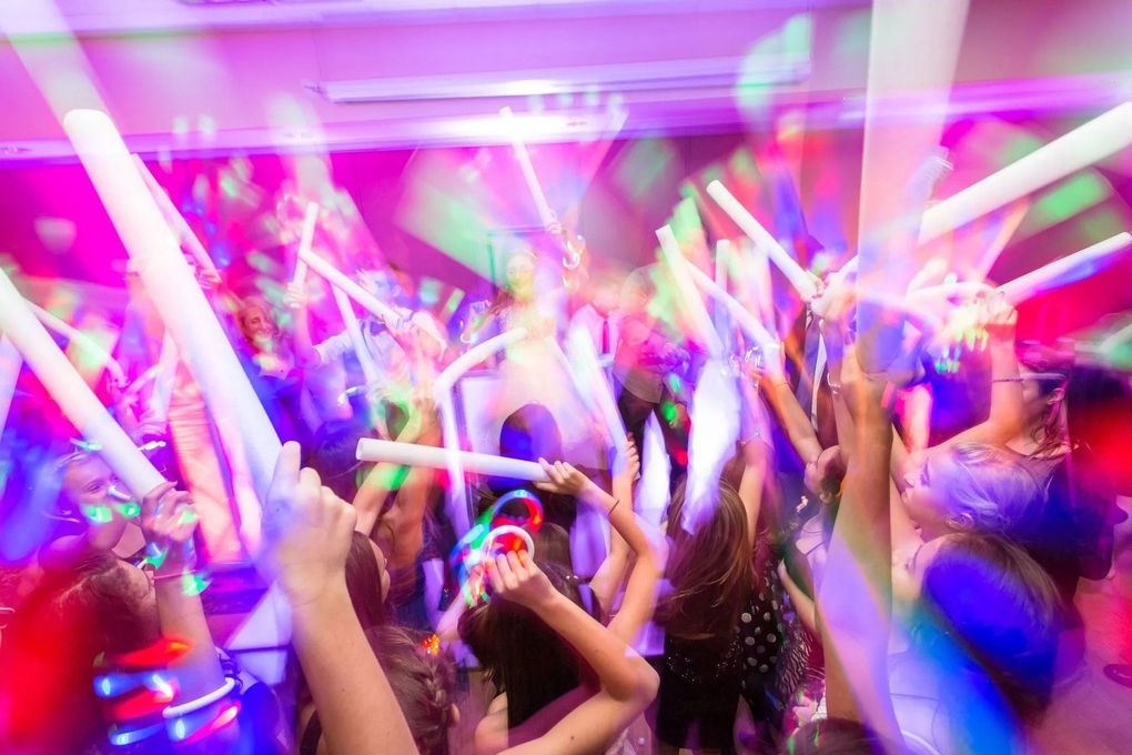 People dancing, waving illuminated foam glow sticks in a colorful, blurred party setting.