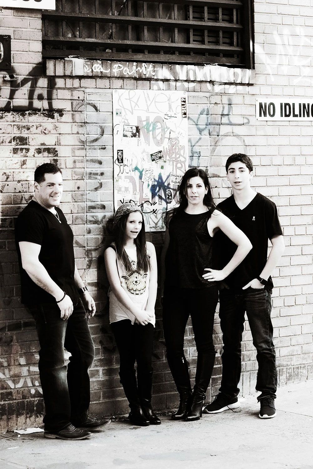 Four people standing against a brick wall with graffiti. They wear dark clothing, looking towards the camera.