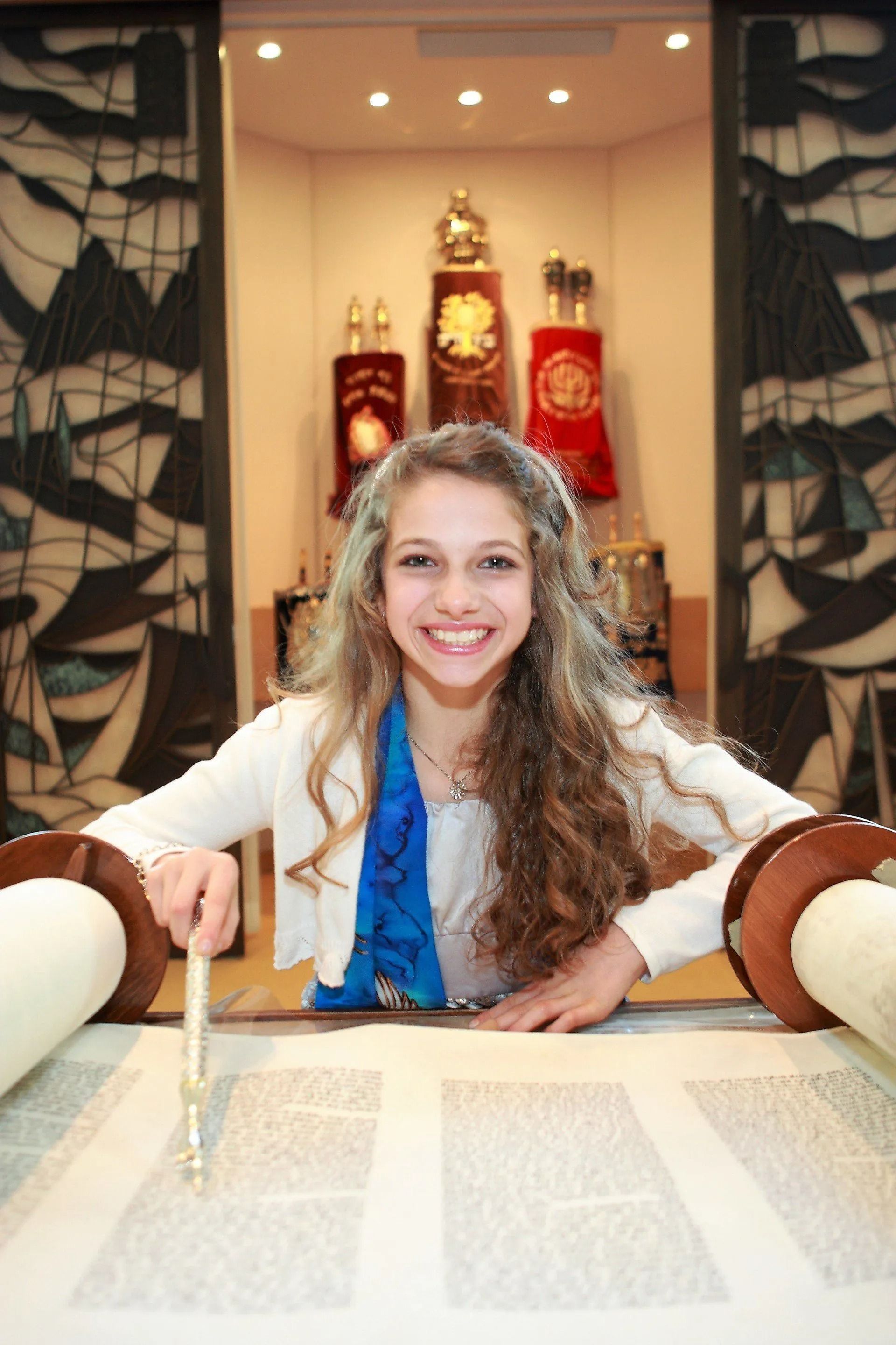 Young person smiling, reading Torah scroll in synagogue. Blue scarf, white jacket.