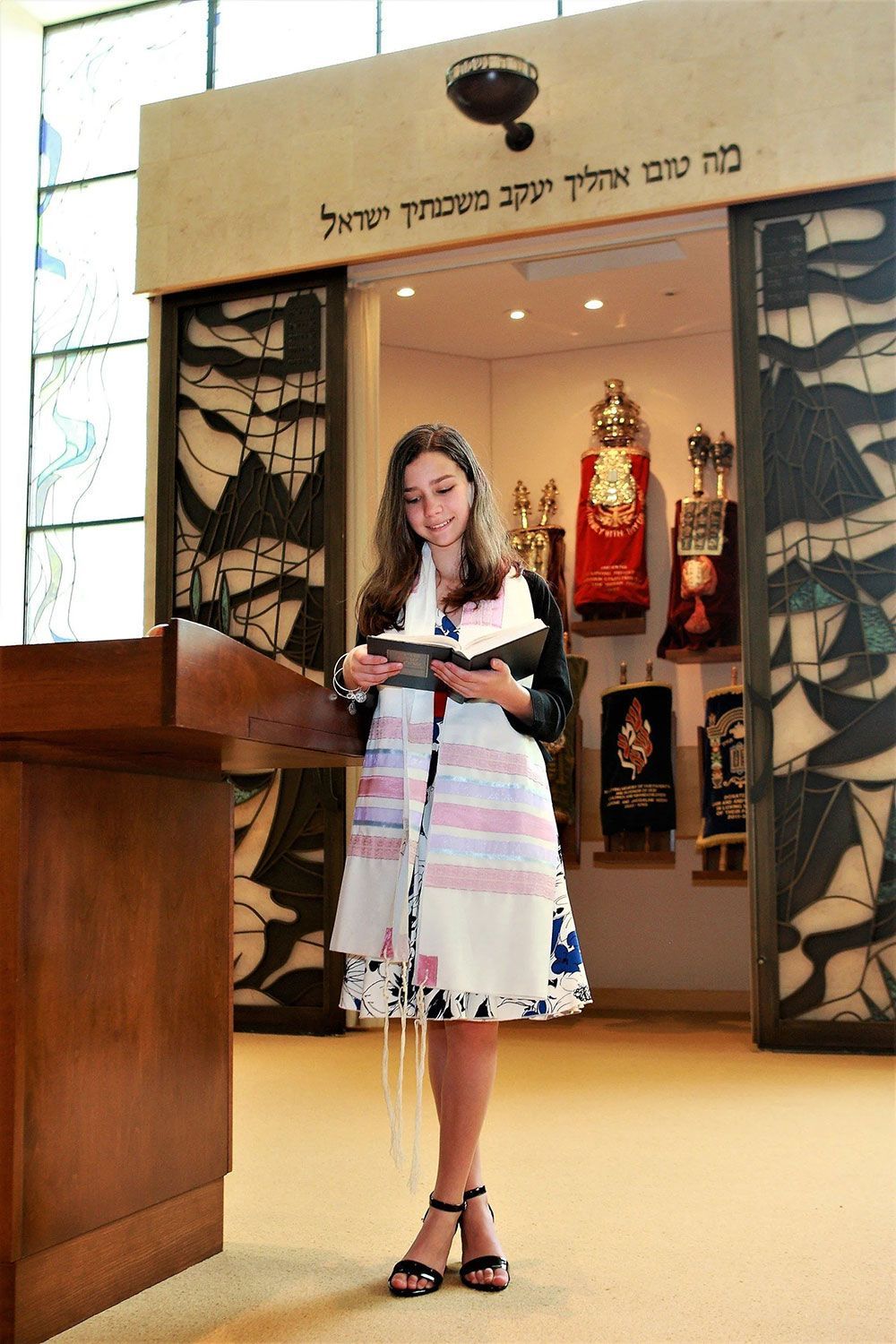 Girl in prayer shawl reads in synagogue, with Torah scrolls visible behind her.