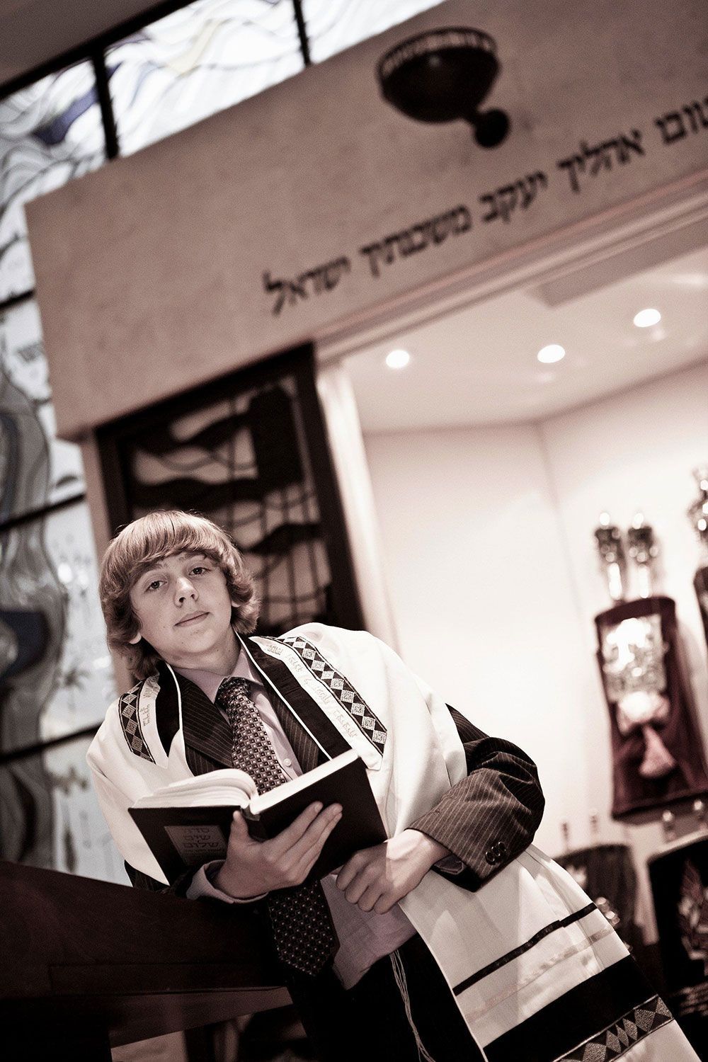 Teen boy in prayer shawl holding a book, standing near a doorway with Hebrew text.
