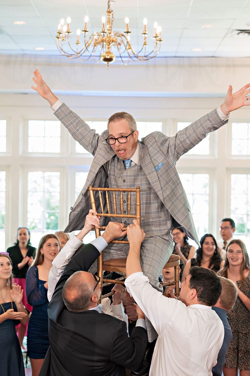 Man in a checkered suit on a chair held high by several people, arms raised, celebratory mood.