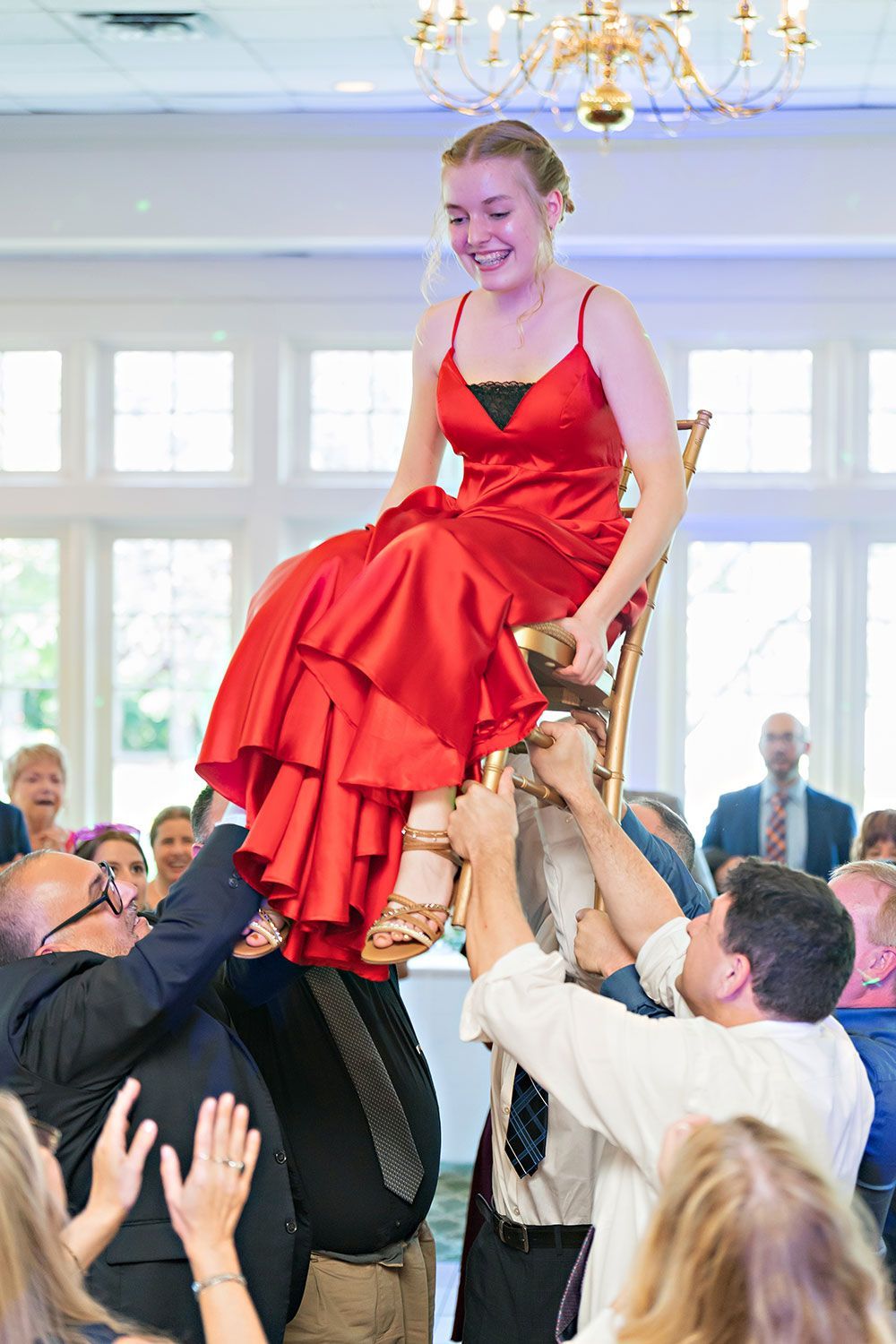 Woman in red dress lifted on a chair by two men at an event, surrounded by people.