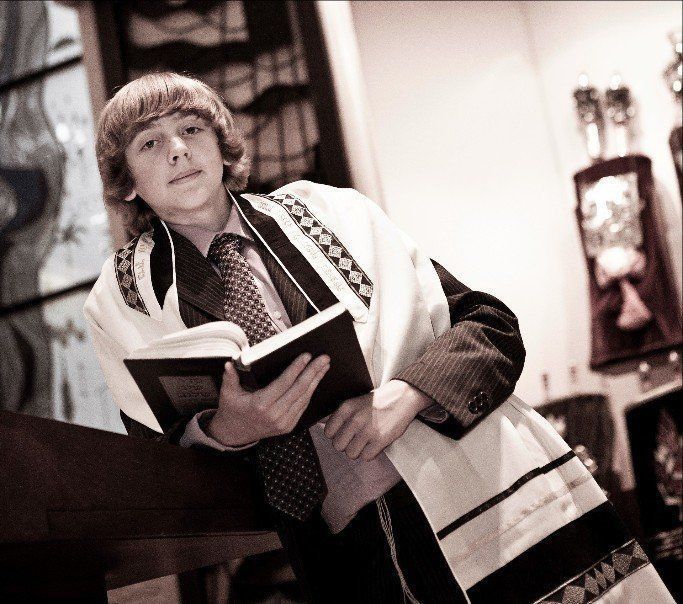 Young person in prayer shawl holding a book, leaning on a dark surface, in a sepia-toned room.