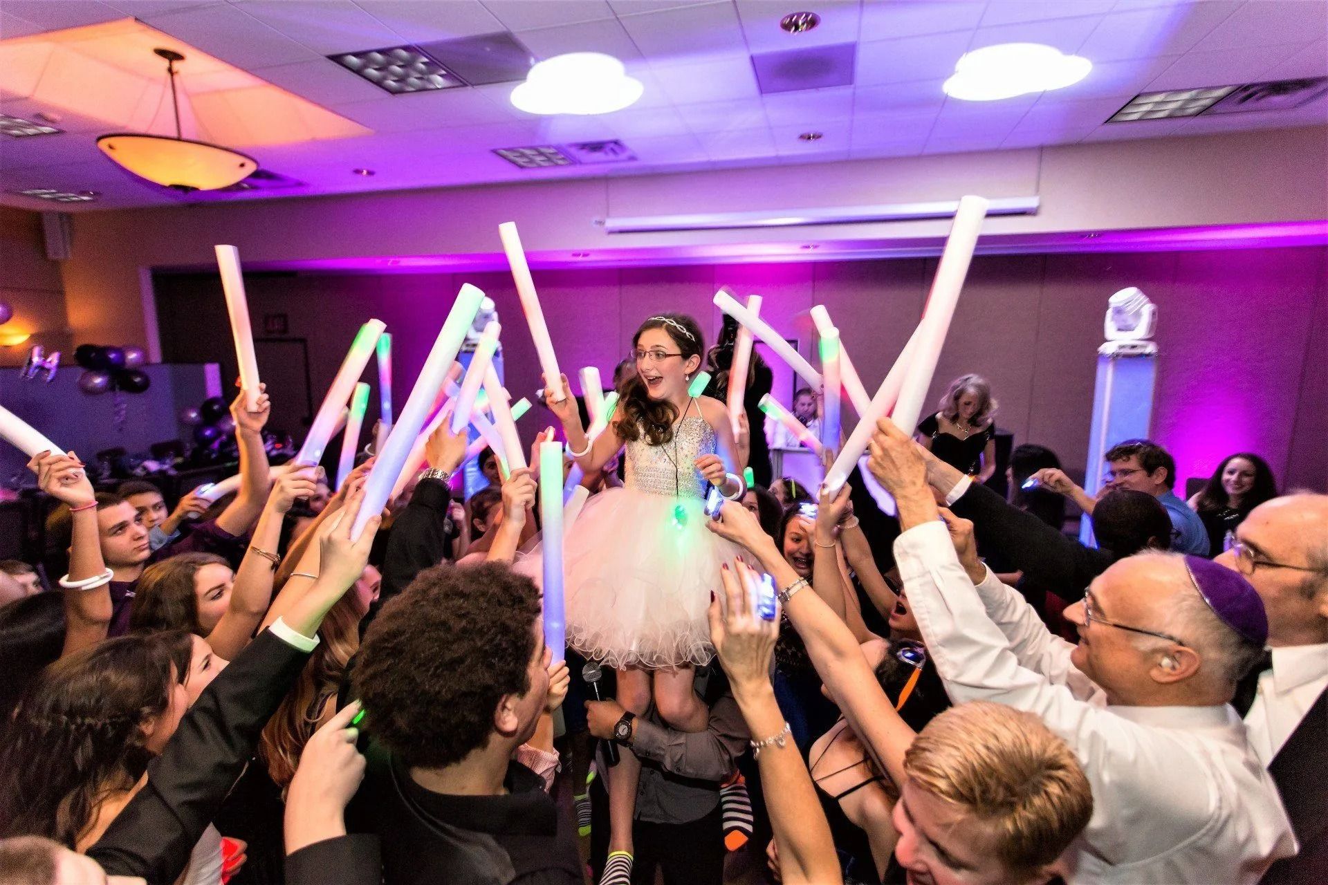 Girl on shoulders at party, surrounded by people holding glow sticks. Purple and white lighting.