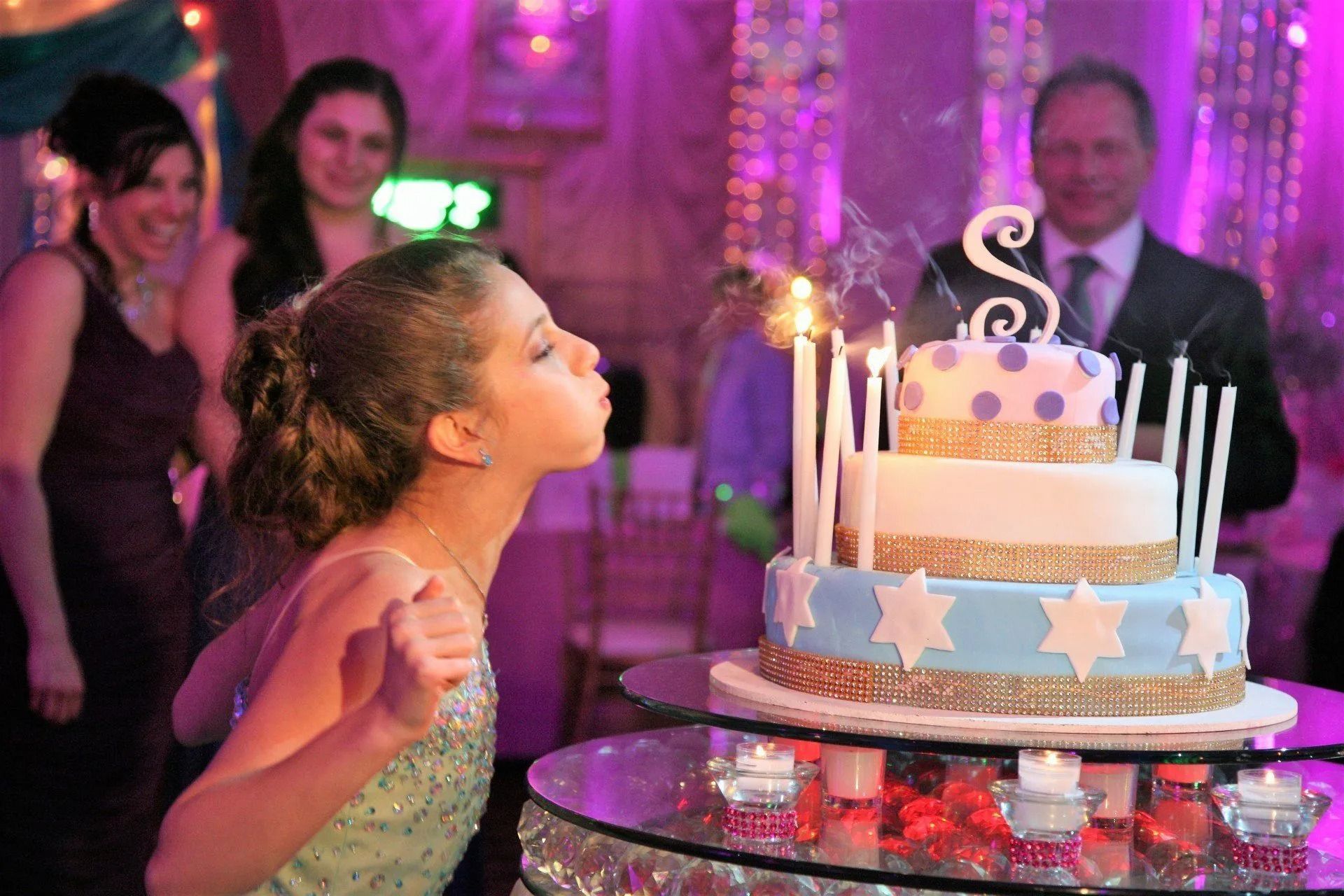 Girl blowing out candles on a tiered birthday cake at a party; purple and white decor, guests watching.