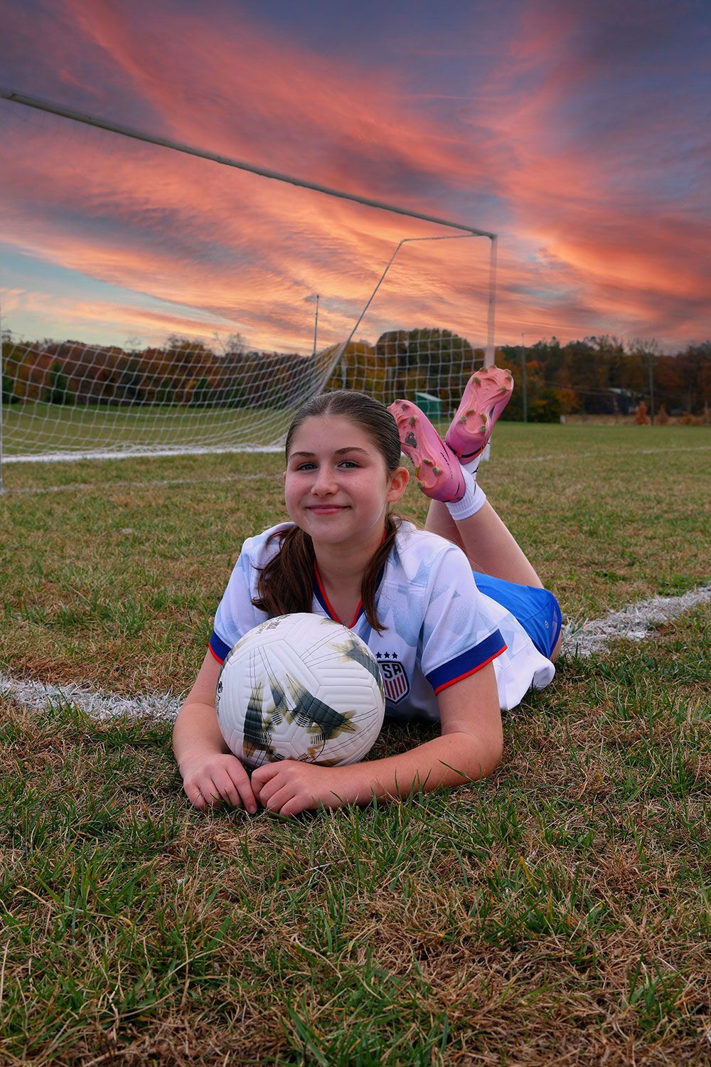 Soccer player lying on grass with ball, goal in background, sunset sky.
