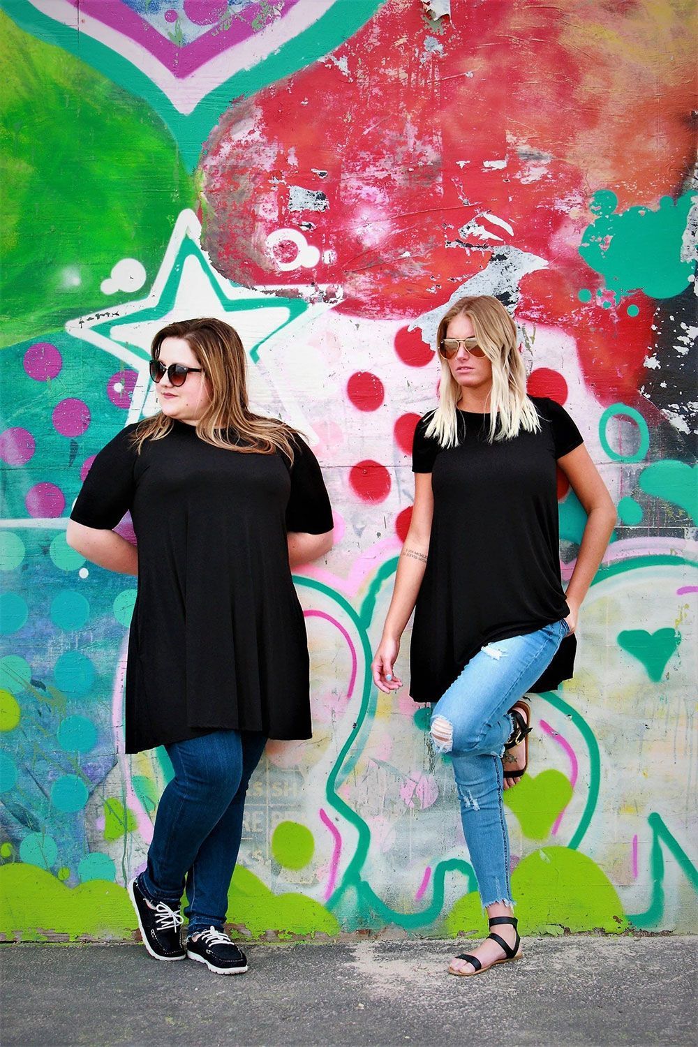 Two women pose by colorful graffiti. They wear black tops, jeans, and sunglasses.