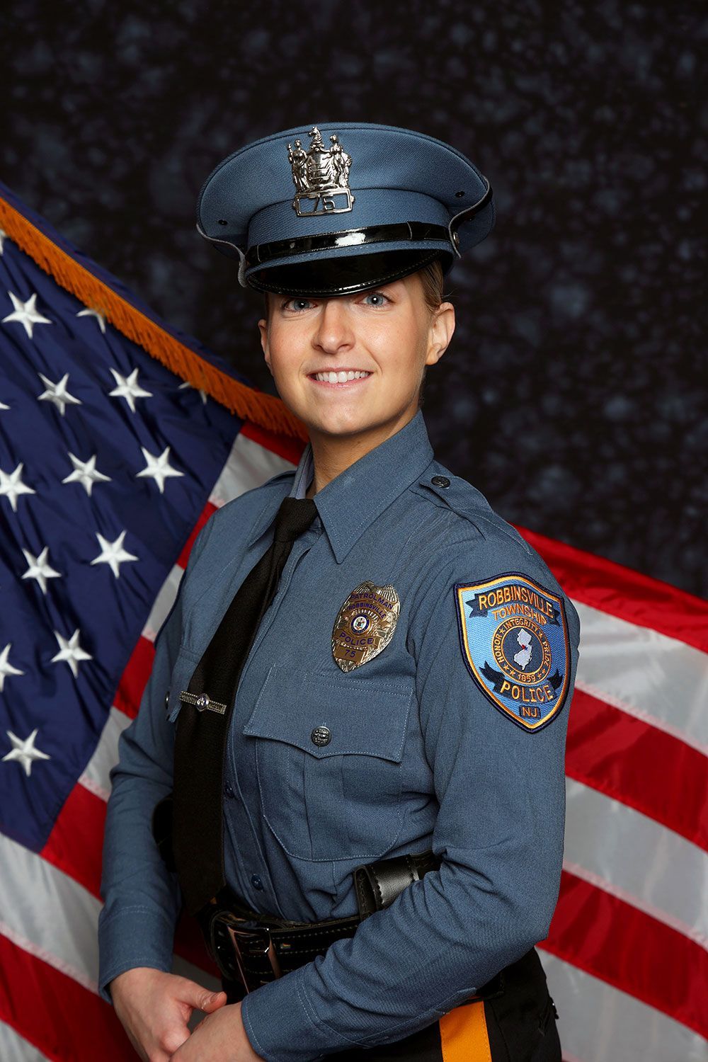 Police officer in blue uniform, smiling, in front of American flag, badge visible.