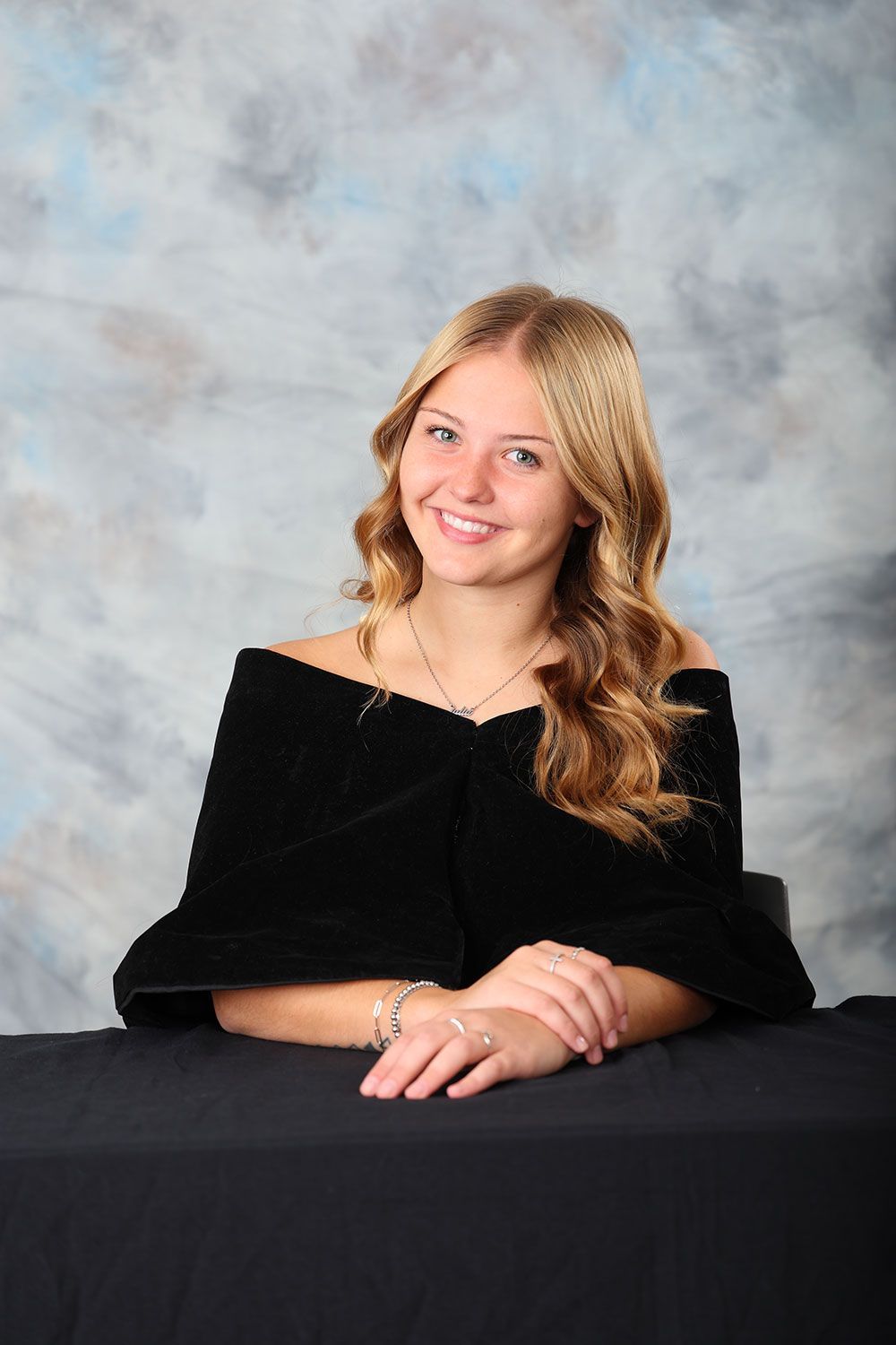 Woman with long blonde hair, smiling, wearing a black off-the-shoulder top, posing in front of a blue and gray background.