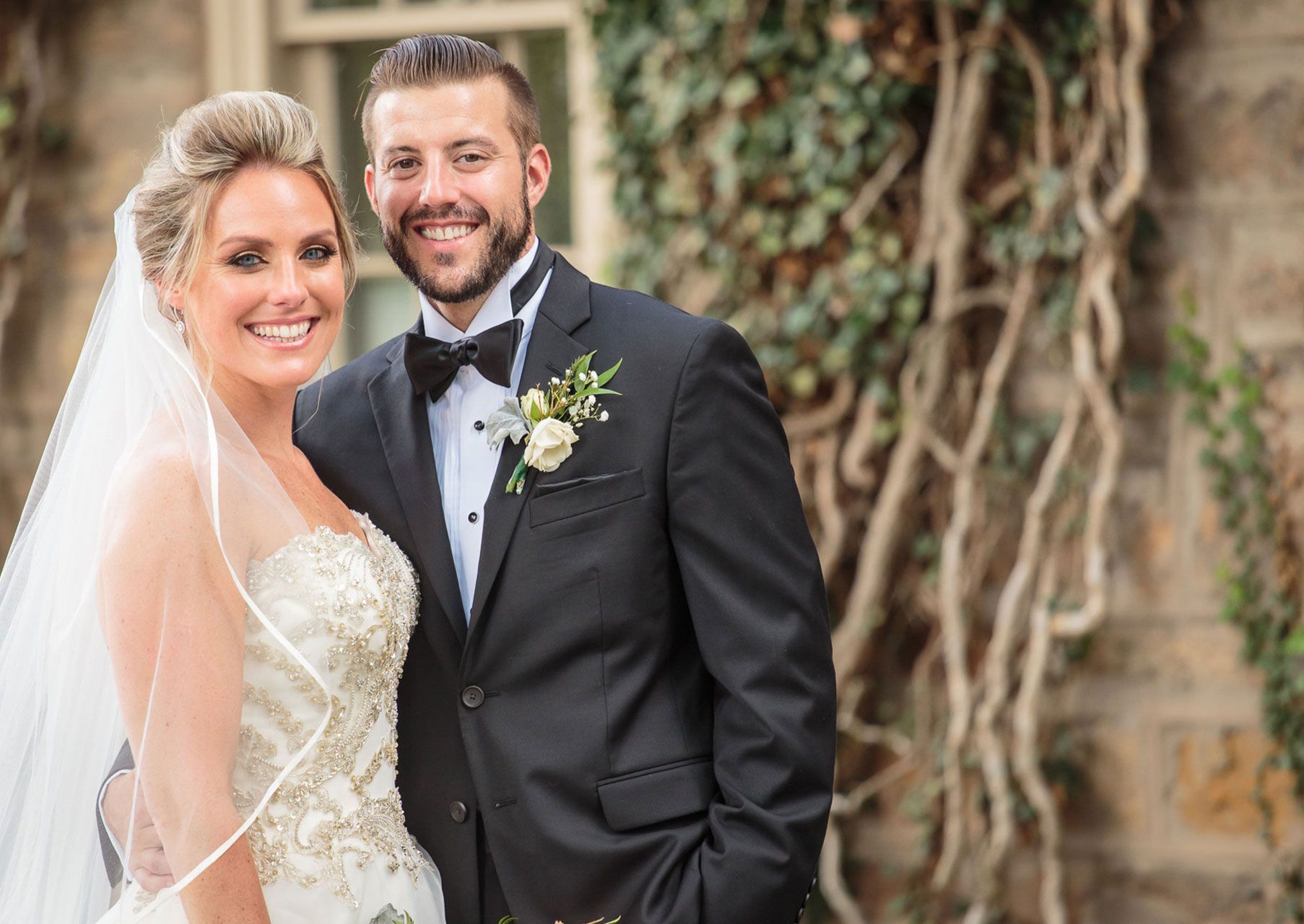 Bride and groom pose outdoors, smiling. The bride wears a strapless gown, the groom a tuxedo, next to ivy-covered stone.