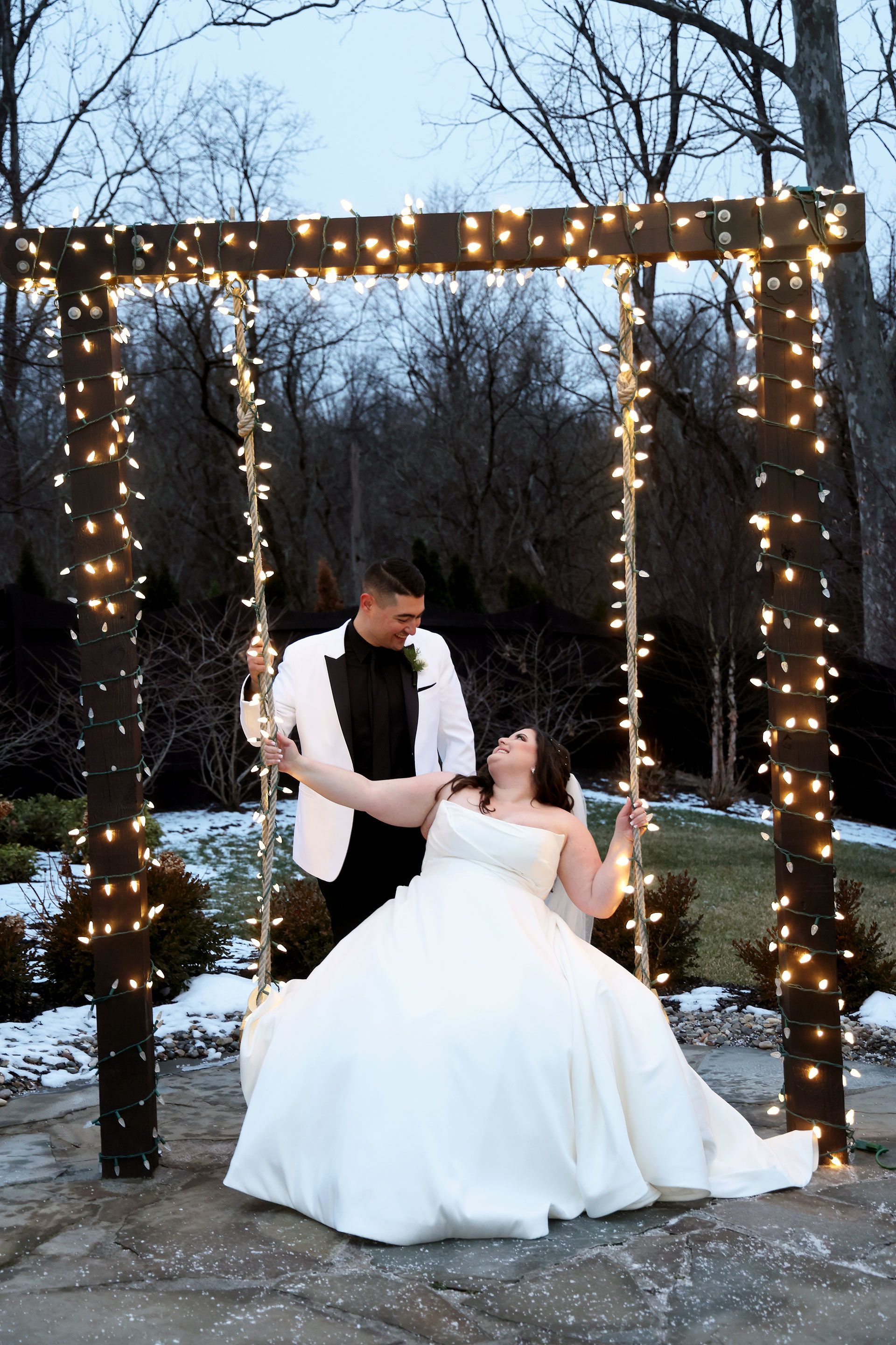 Couple on a swing, decorated with lights. The bride wears a white gown; the groom, a white tuxedo jacket. Outdoor setting.