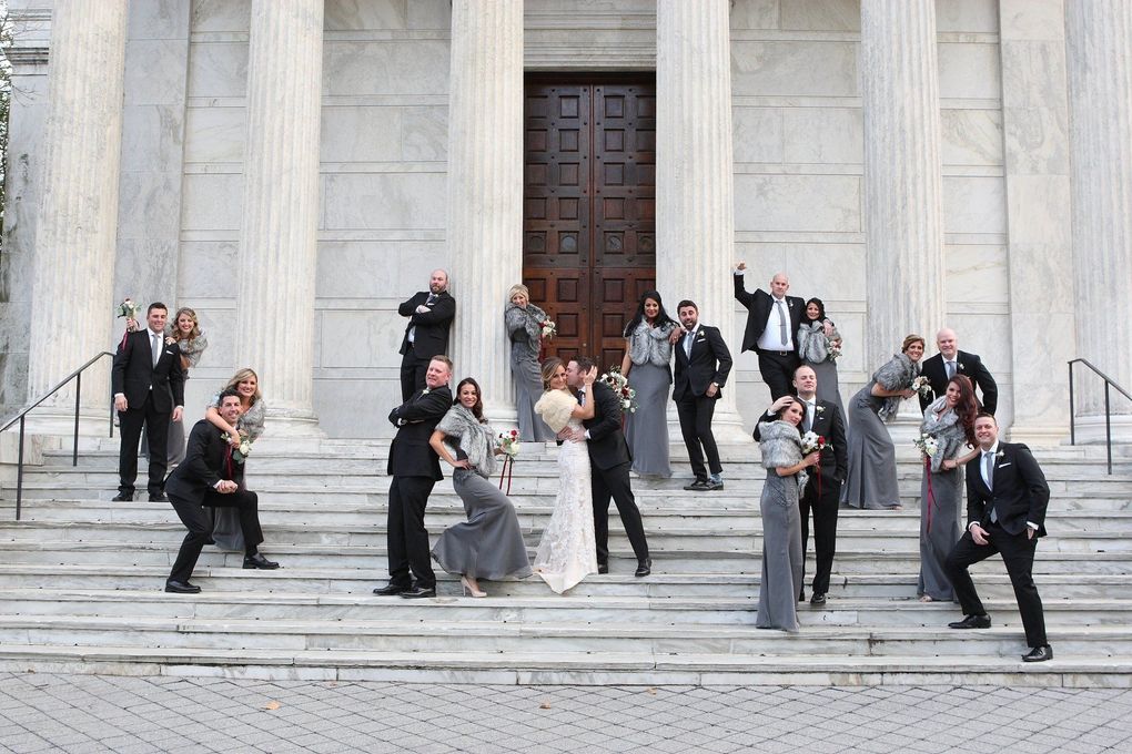 Wedding party posing on steps of a white building with columns. Bride and groom in center, others in formal attire.