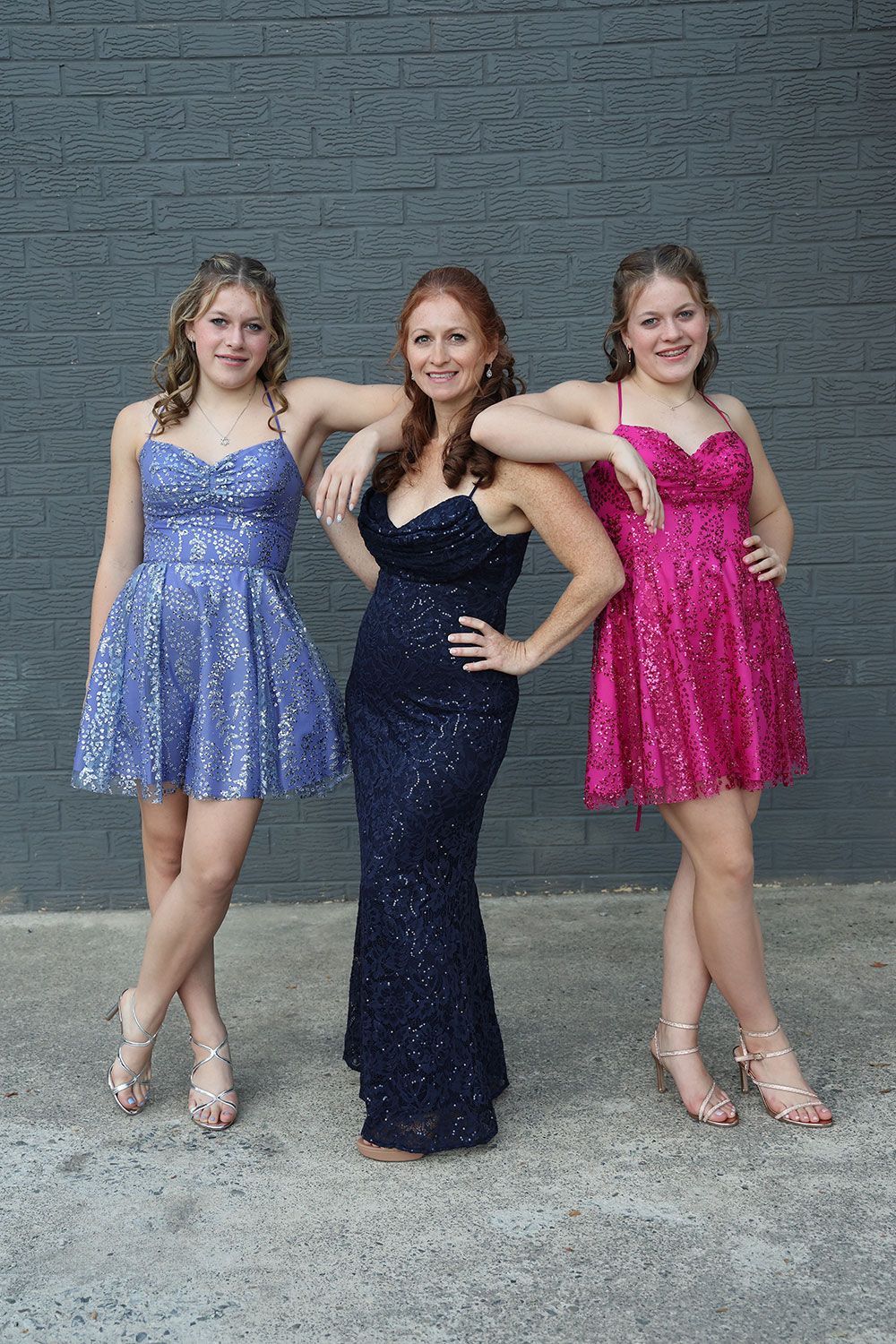 Three people posing against a gray wall. Two in sequined dresses, one blue, one pink, and one woman in a sequined navy gown.