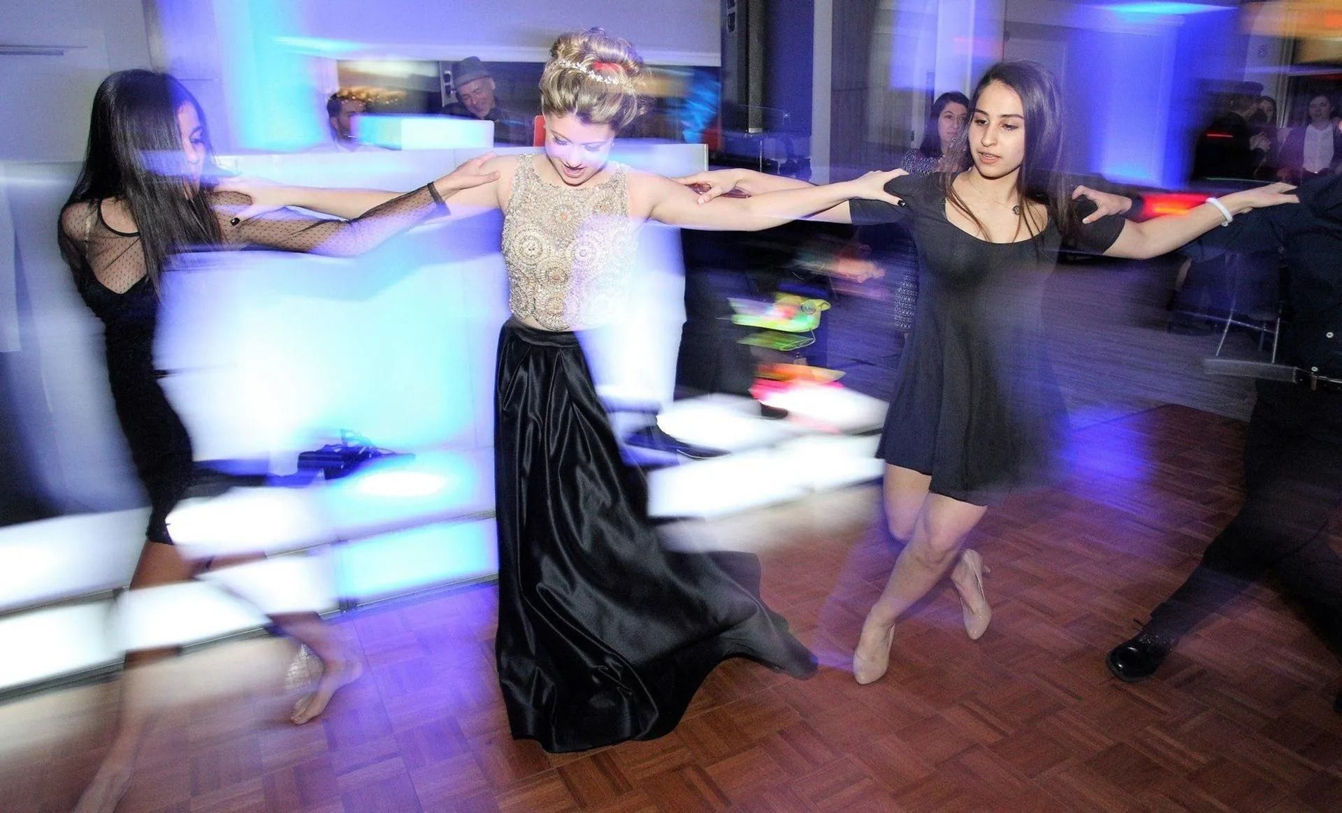 Three people dancing in a circle on a wooden floor, blurred by motion; one wears a formal gown.