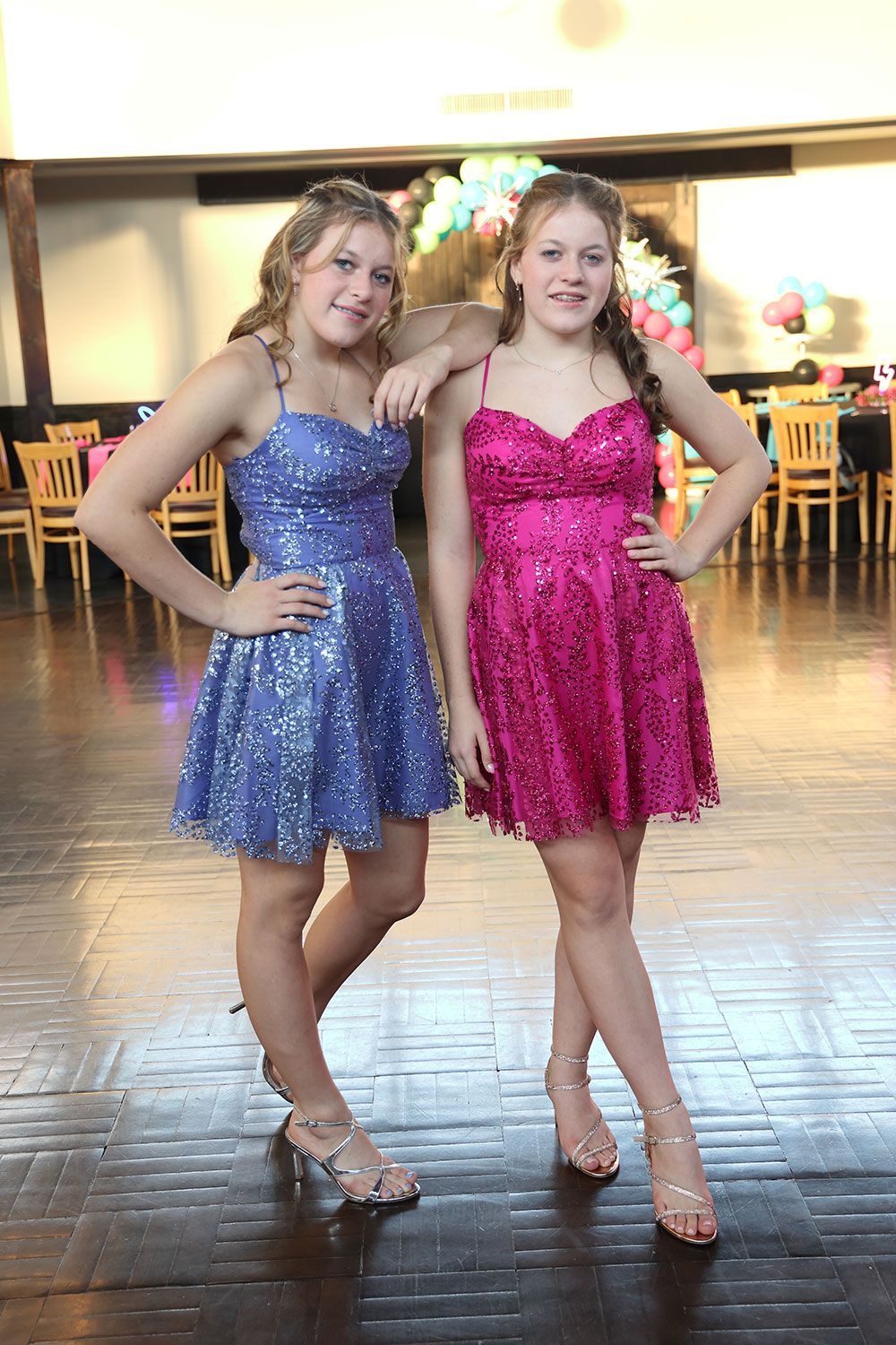 Two young women in sparkly dresses pose at a dance; one in blue, one in pink.
