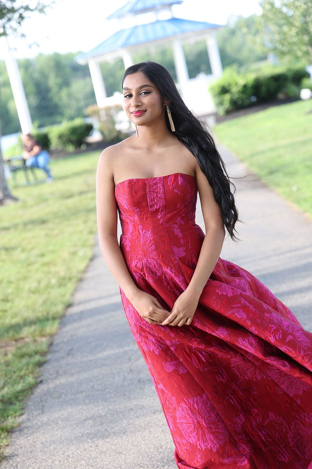 Woman in red strapless gown stands on path, hair flowing. Outdoors, with a gazebo and trees in background.