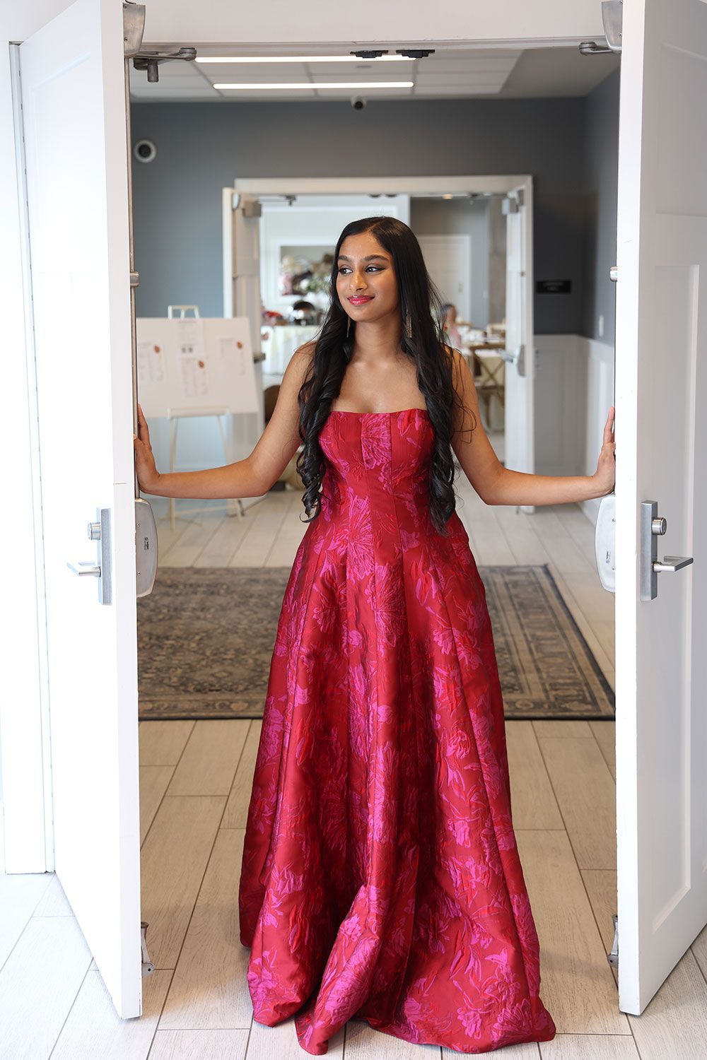 Woman in a red sparkly ball gown stands in doorway, looking away, arms extended.