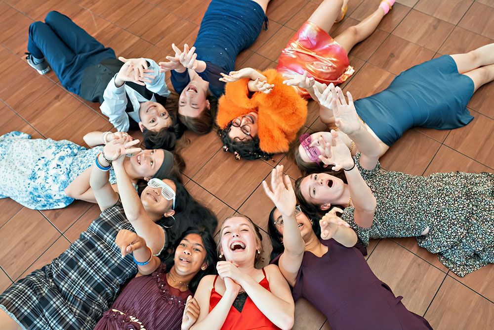 Ten people lying in a circle on a wooden floor, looking up and smiling.