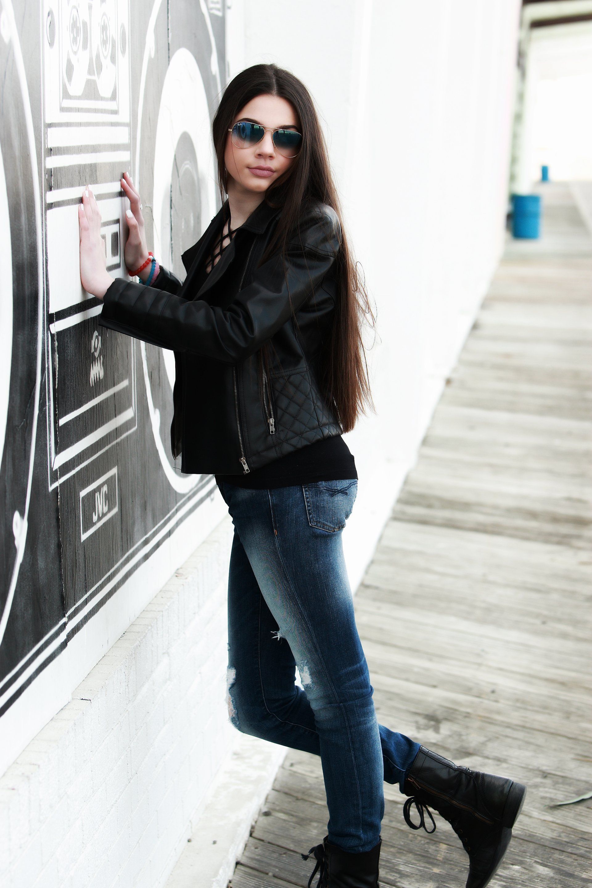 Woman in sunglasses and leather jacket leans against a wall with graffiti, wearing jeans and boots.
