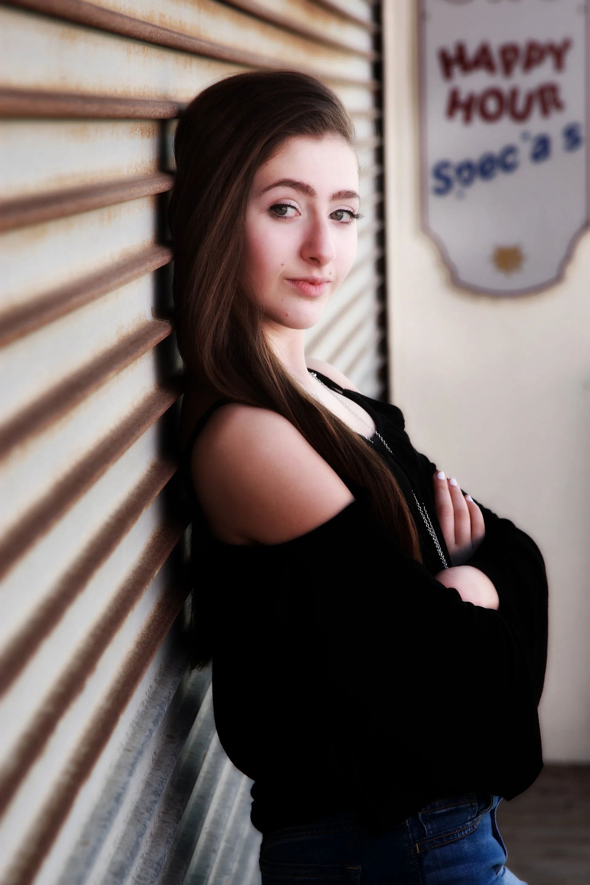 Woman leans against a weathered metal wall, arms crossed, looking at the camera.