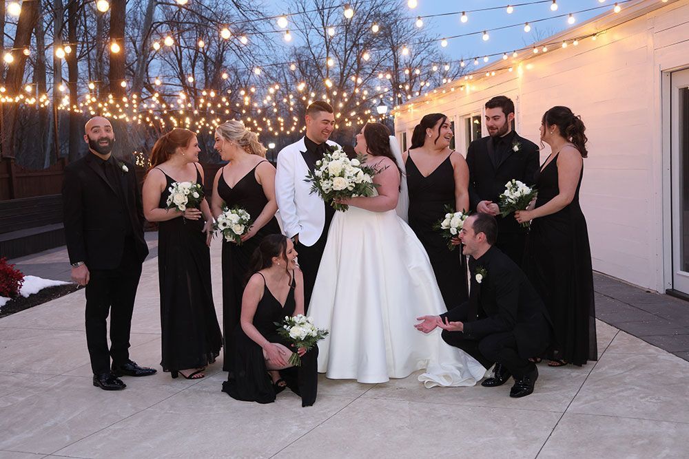 Wedding party: Bride and groom with bridesmaids and groomsmen outdoors, under string lights.