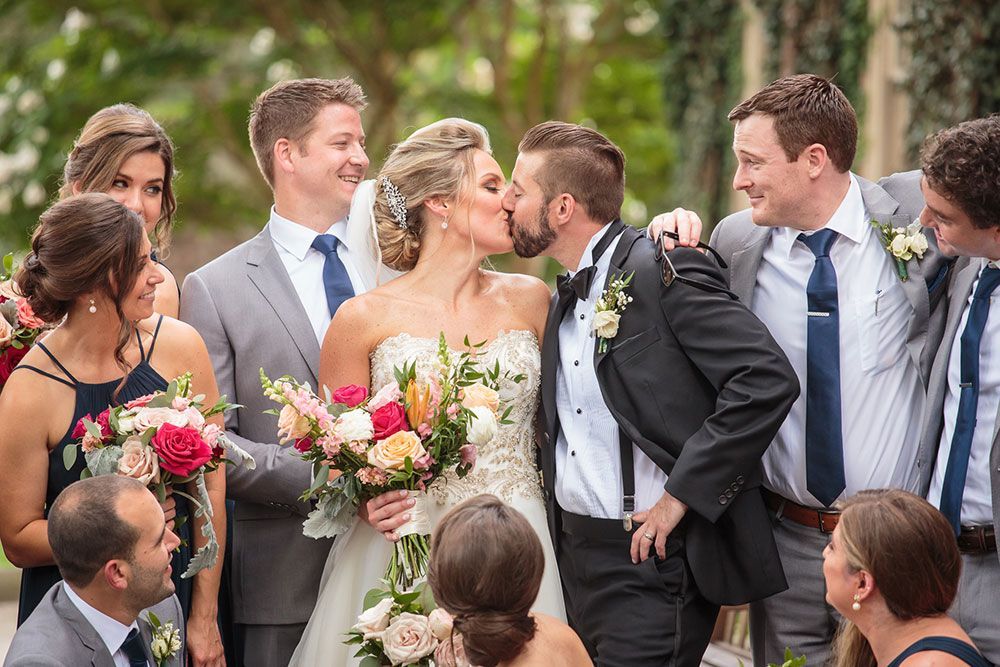 Bride and groom kissing, surrounded by wedding party. Outdoors with greenery, bright colors.