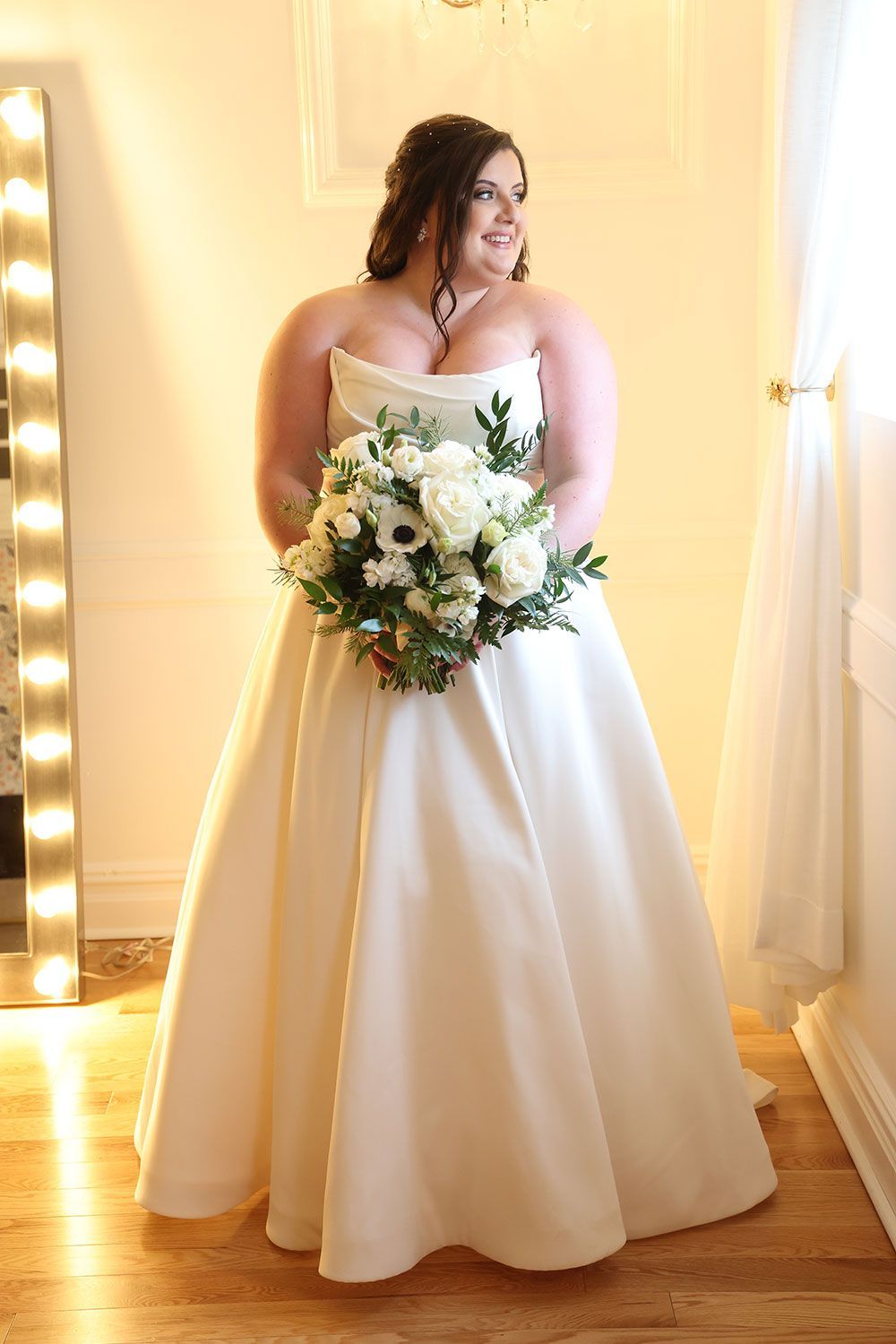 Bride in white strapless dress holding a bouquet, standing by a window.