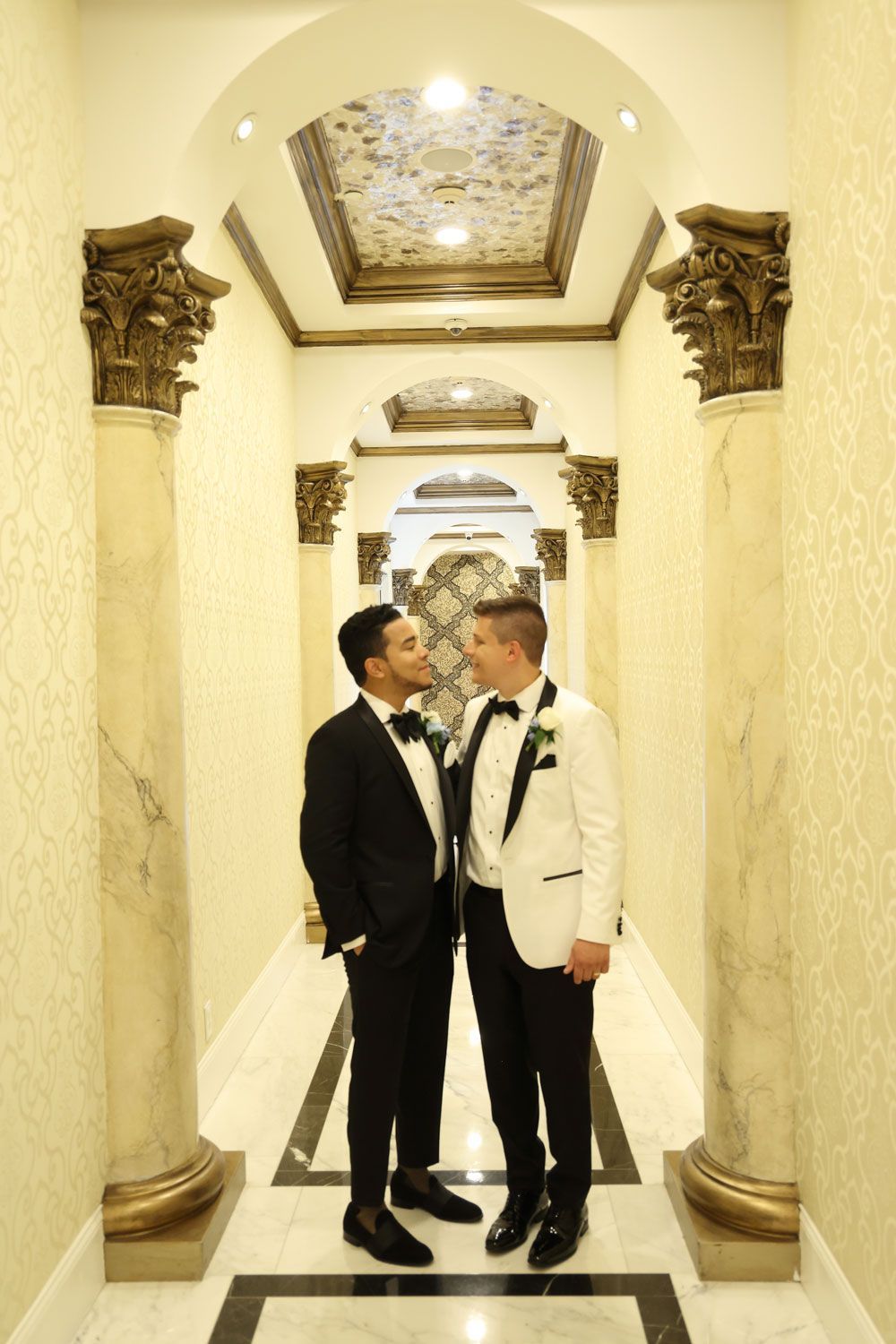 Two men in tuxedos, facing each other, in a hallway with ornate pillars.
